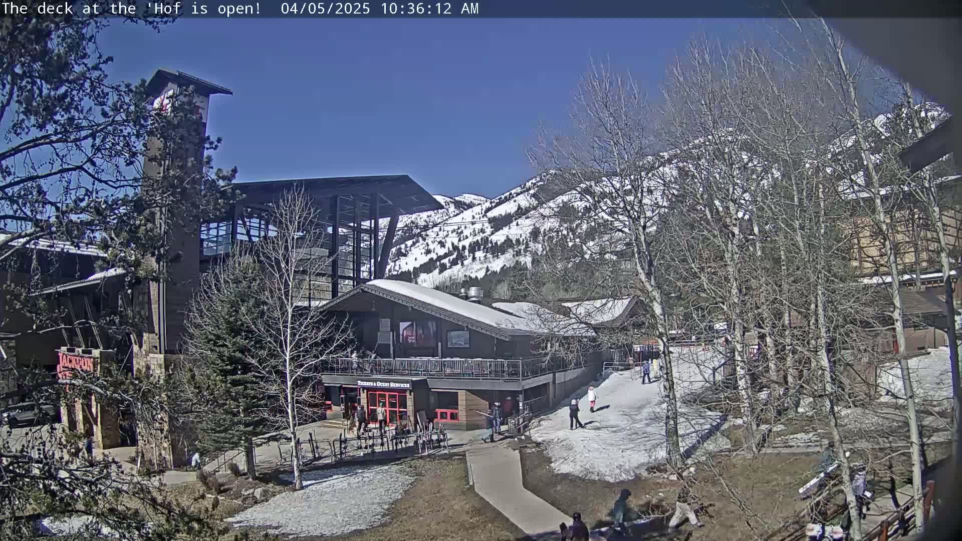 A snow-dusted mountainside is visible behind a modern building complex with a deck,  under a clear blue sky.