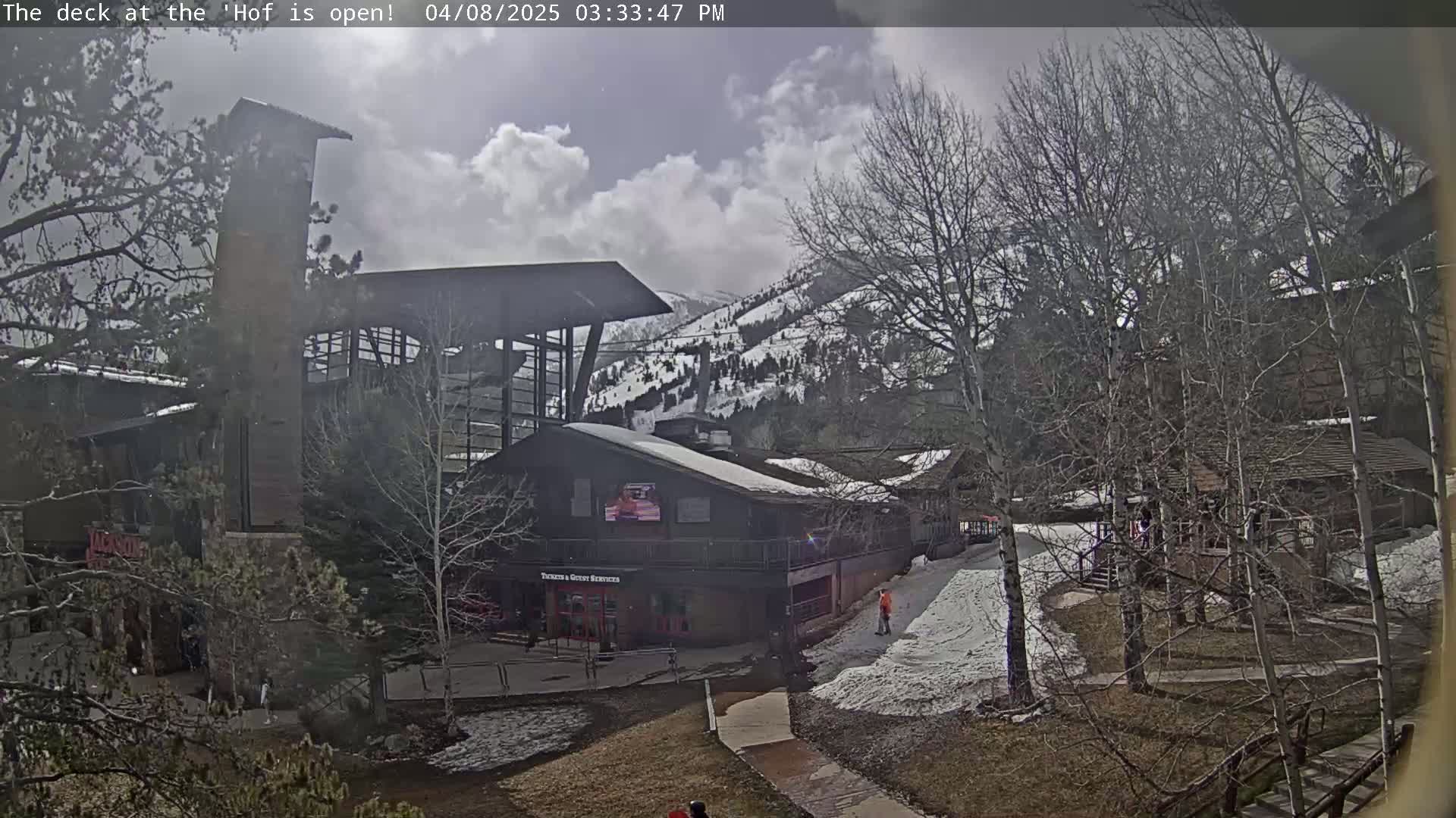 A partially cloudy day reveals a snow-dusted mountainside behind several buildings and walkways, with bare trees scattered throughout the scene.