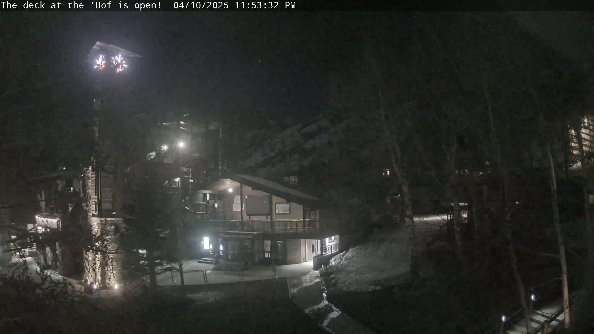 A nighttime view of a snow-covered lodge and surrounding trees, illuminated by lights, with fireworks visible in the distance.