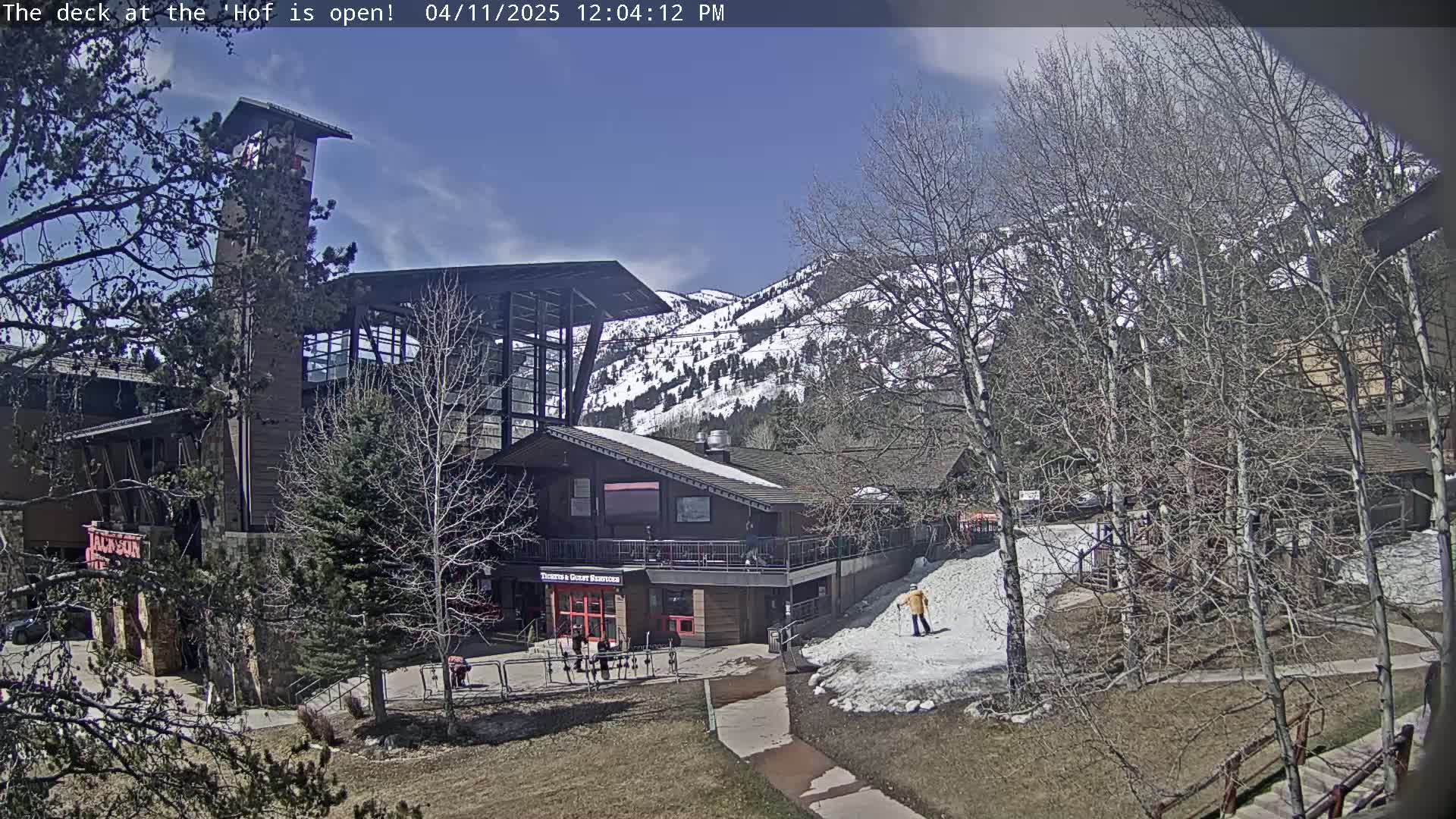 A lodge-style building with a deck sits partially surrounded by bare trees and patches of snow on a sunny day with mountains in the background.