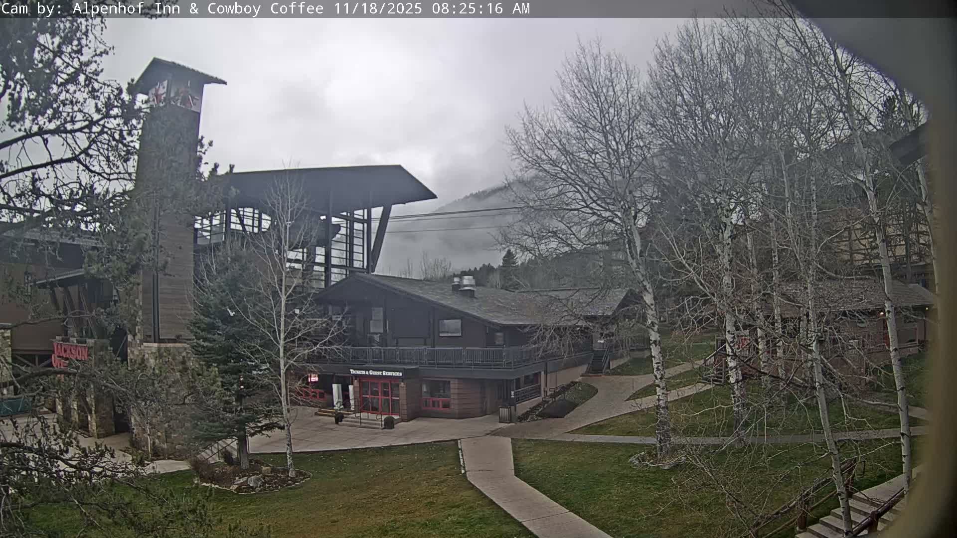 A nighttime, low-light view shows a two-story building with a balcony, illuminated by several lights, surrounded by dark trees.
