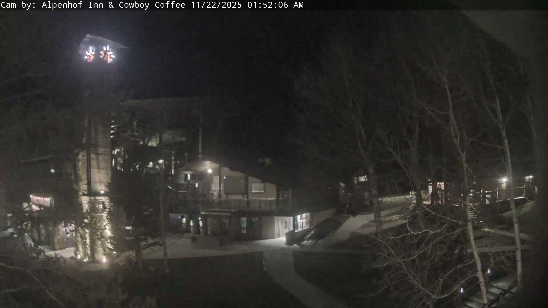 A clear, dark night reveals an outdoor scene with multiple illuminated buildings, including a prominent lodge with bright decorative star-shaped lights, surrounded by bare-branched trees and winding paths.