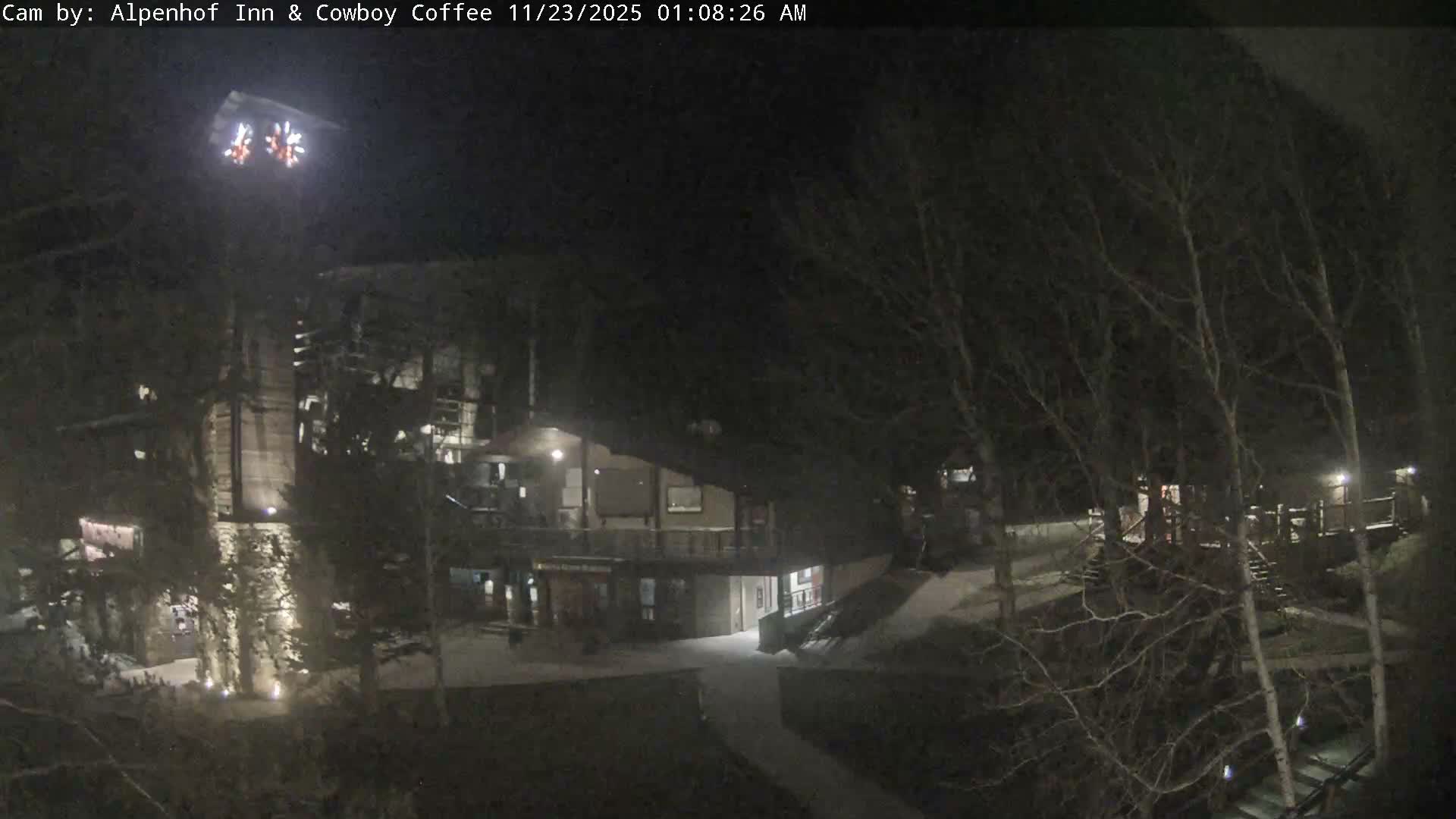 A nighttime view reveals a well-lit lodge or inn adorned with decorative lights above, surrounded by bare-branched trees and illuminated pathways under a clear night sky.