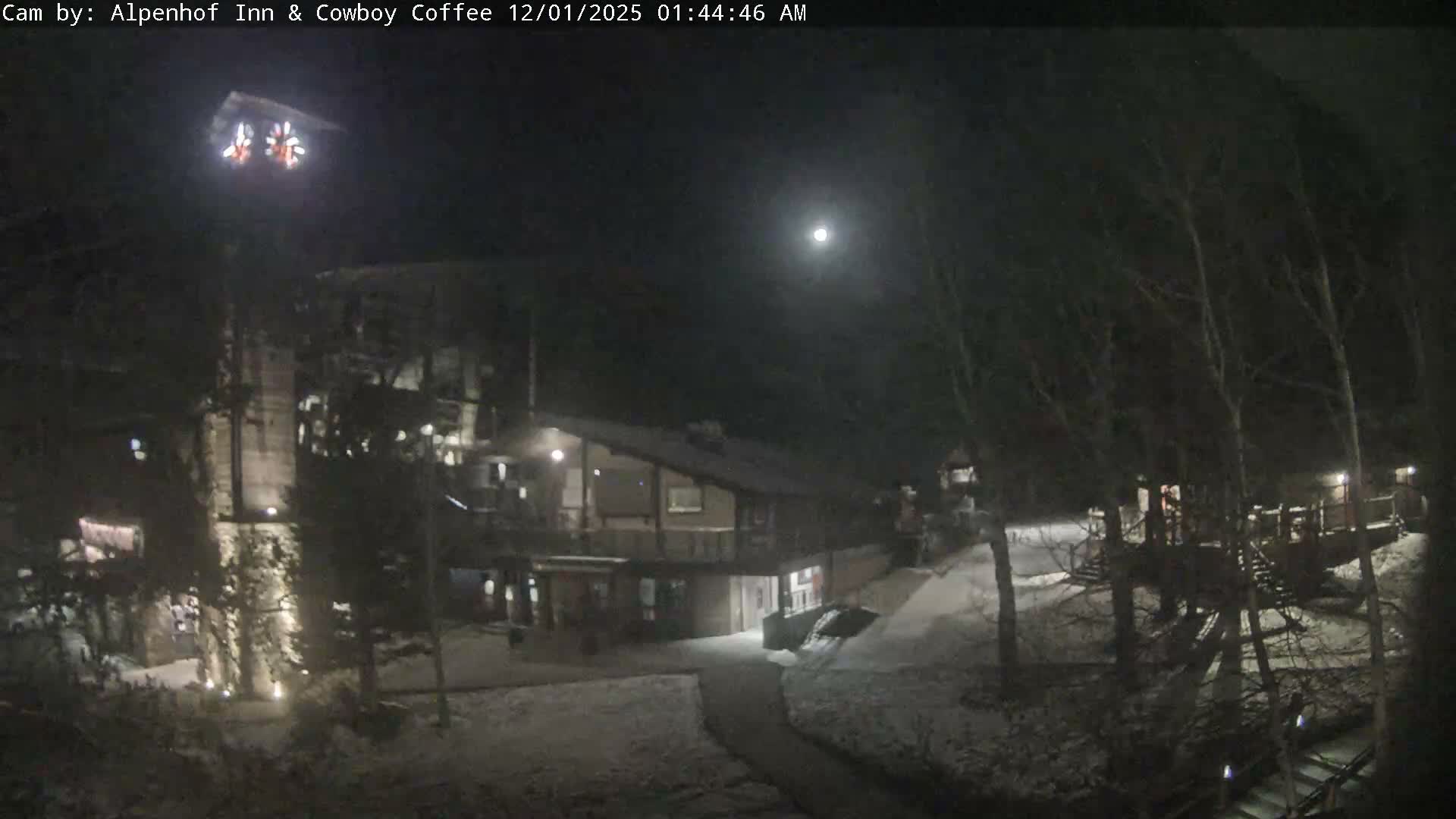 A clear winter night illuminates a snow-covered lodge and surrounding bare trees under a bright moon in the dark sky.