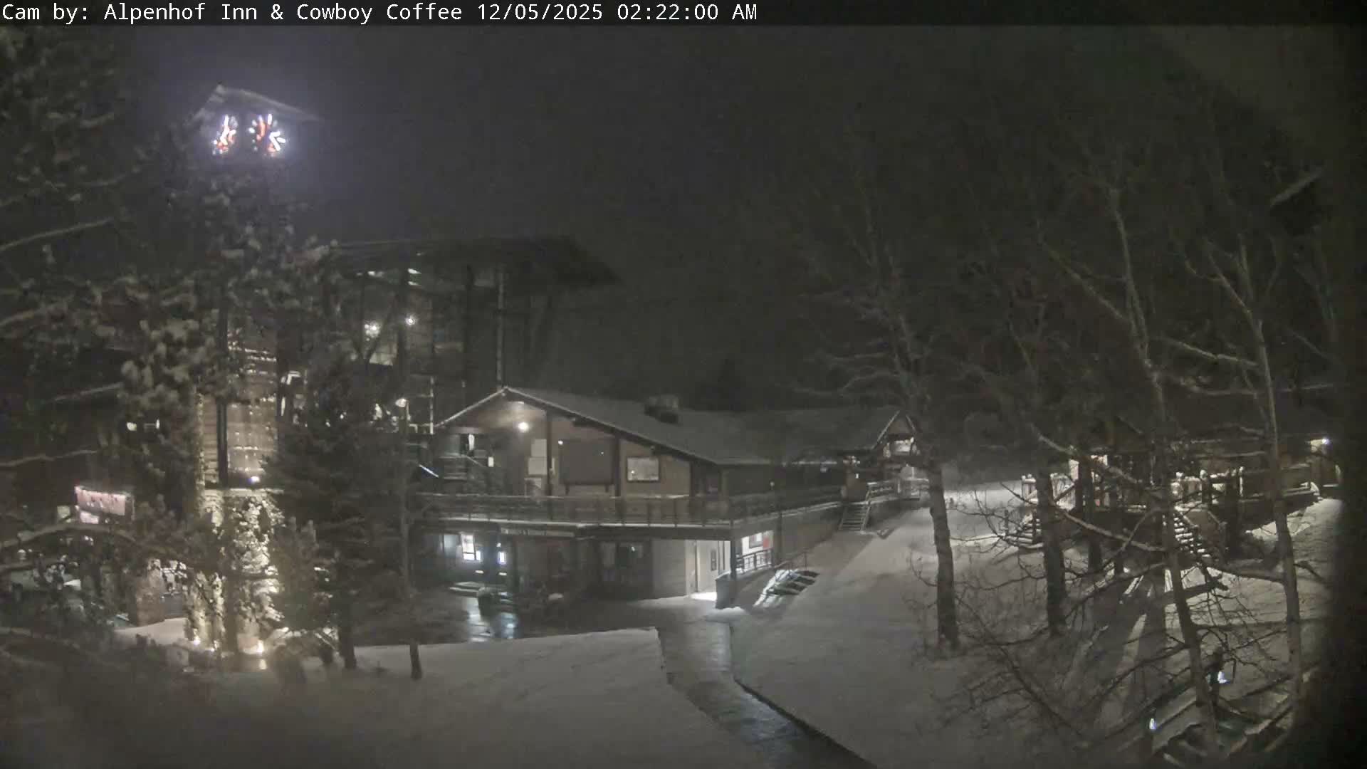 A snowy, winter night reveals a warmly lit lodge or resort, featuring several buildings with snow-covered roofs and grounds, surrounded by trees dusted with snow, under a dark sky with a light snowfall.