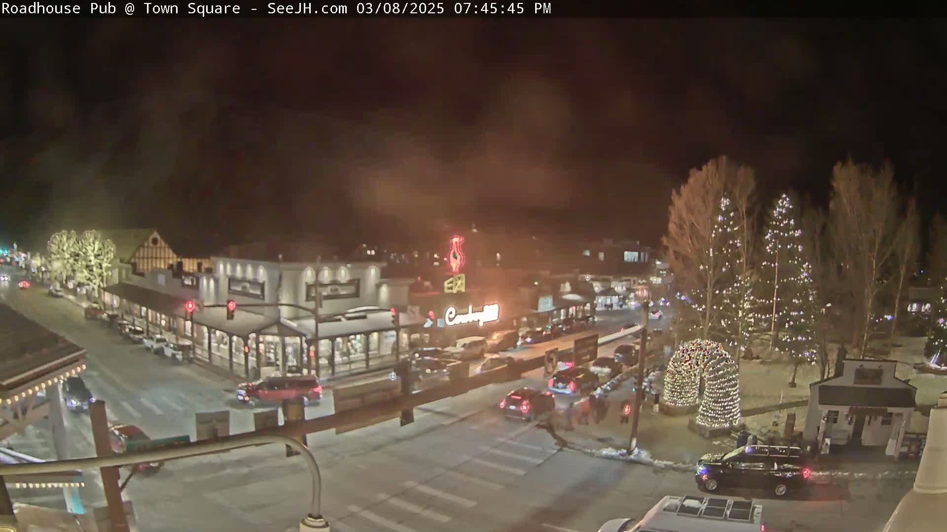 A nighttime view of a snow-dusted town square with buildings, vehicles, and Christmas lights illuminating trees and a decorative arch.