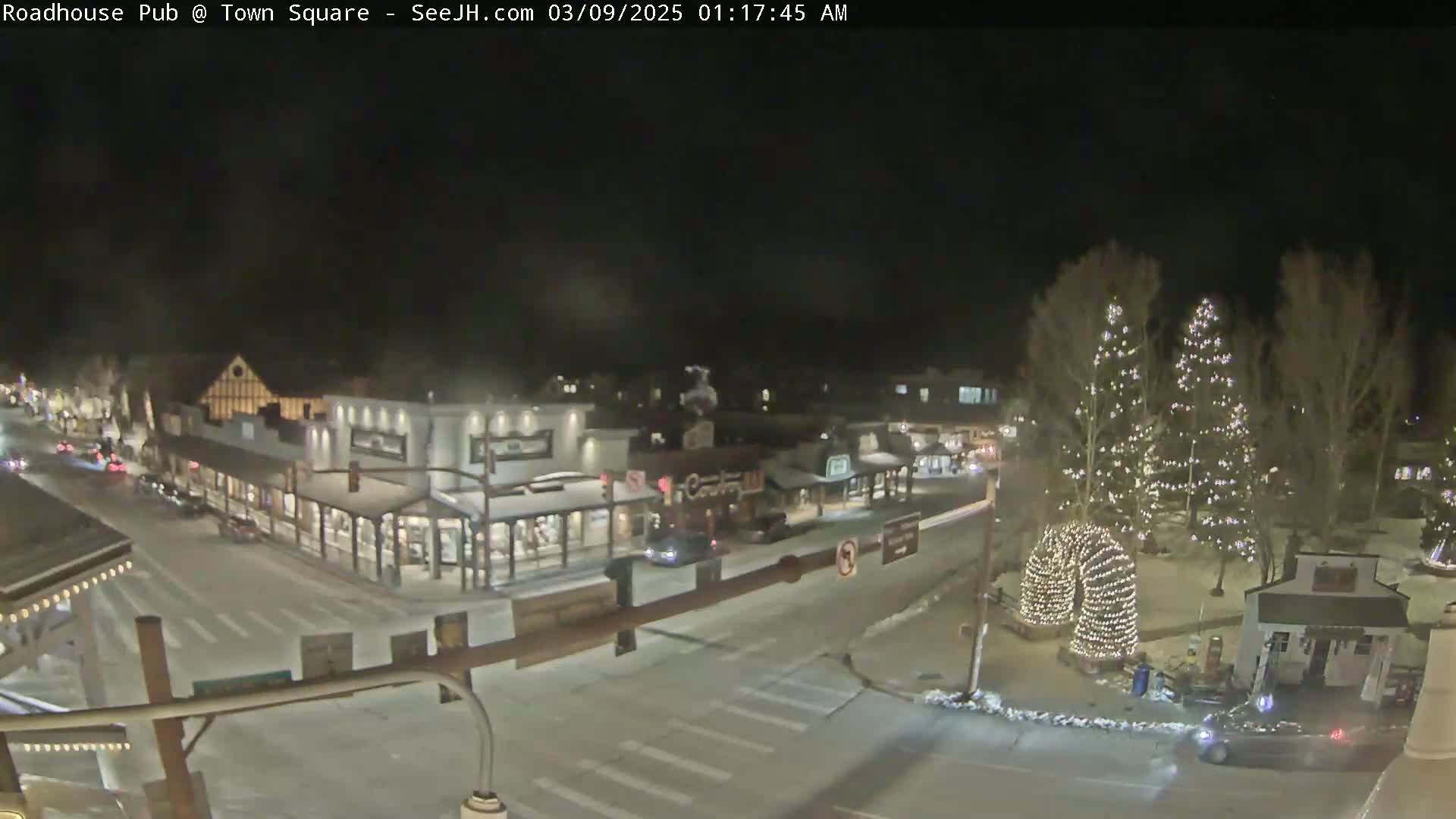 A snow-covered town square at night, illuminated by streetlights and Christmas lights on trees, with cars driving on the roads.