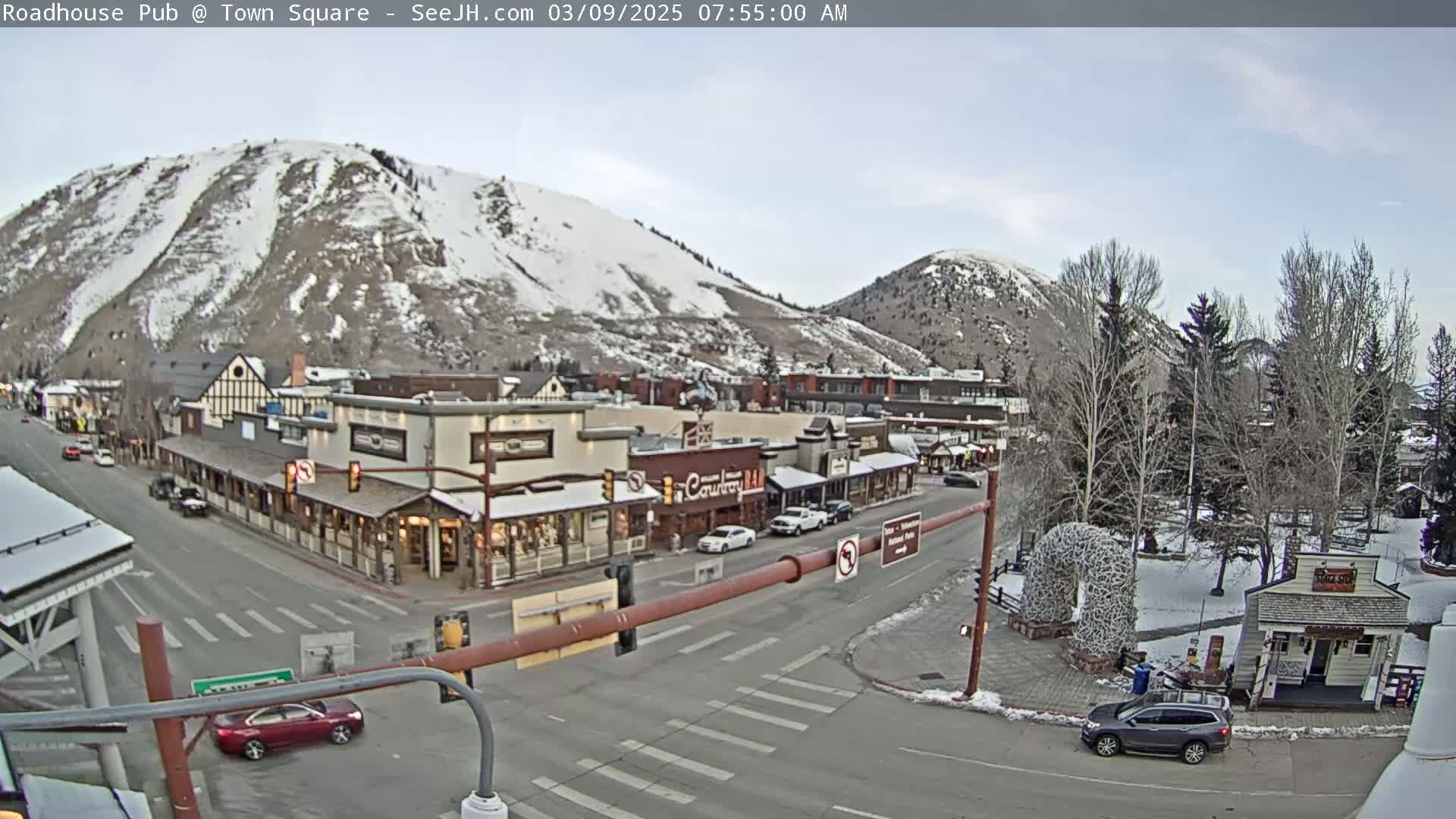 A partly cloudy morning scene shows a snow-dusted mountain overlooking a town square with several buildings and cars, and a few bare trees in a park.
