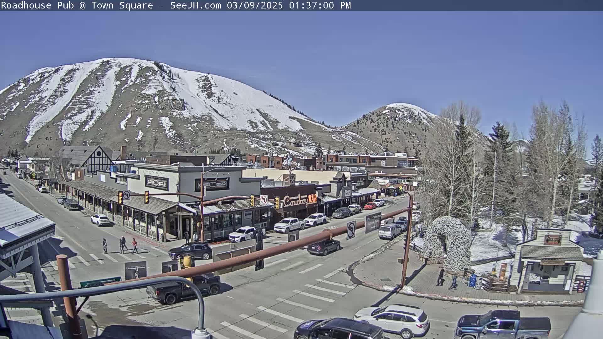 A sunny day reveals a town square with snow-capped mountains in the background, several buildings lining a street filled with parked and moving vehicles, and pedestrians crossing the road.