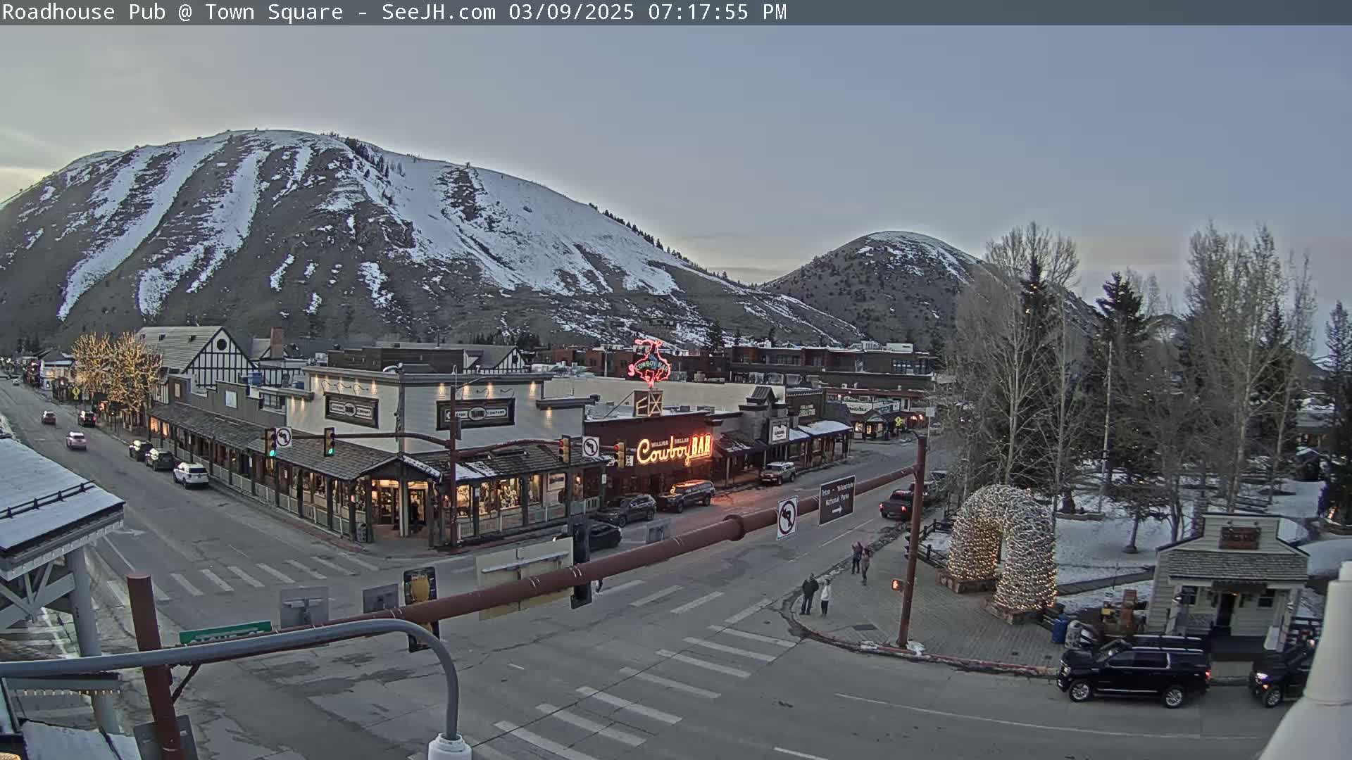 A snow-dusted mountain overlooks a town square with buildings and cars under a twilight sky.