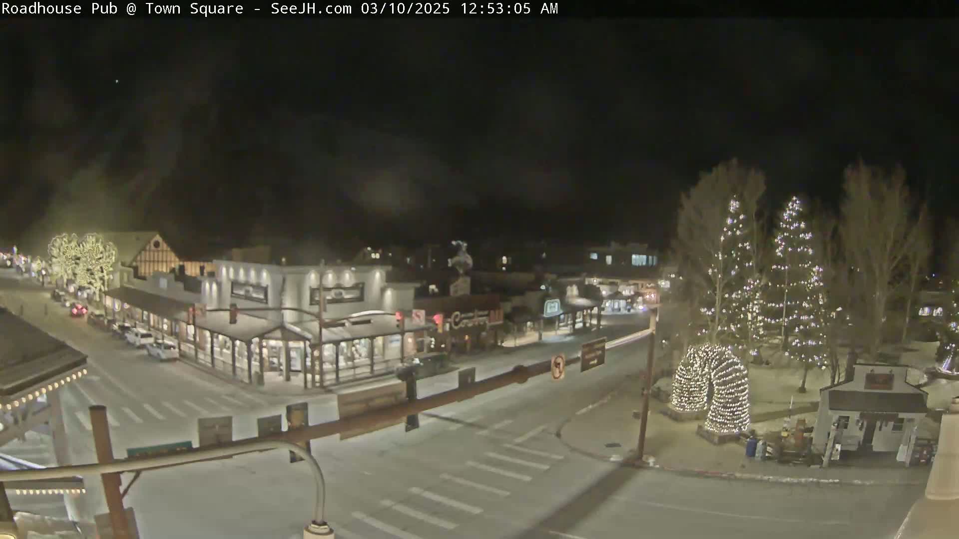 A nighttime view of a snow-covered town square with illuminated Christmas trees and buildings.