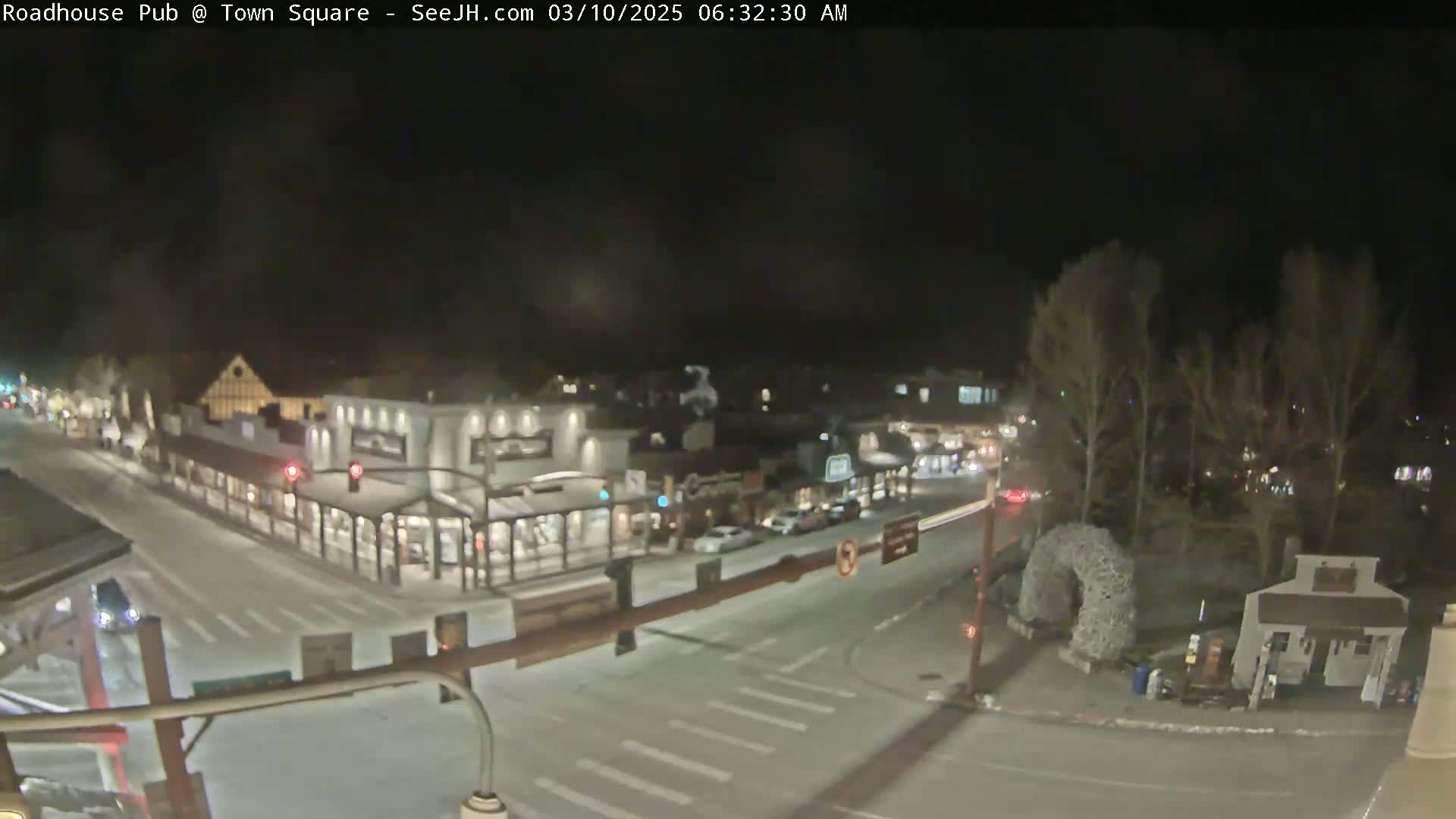 A nighttime view of a snow-covered town square with several buildings and cars, under a dark sky.