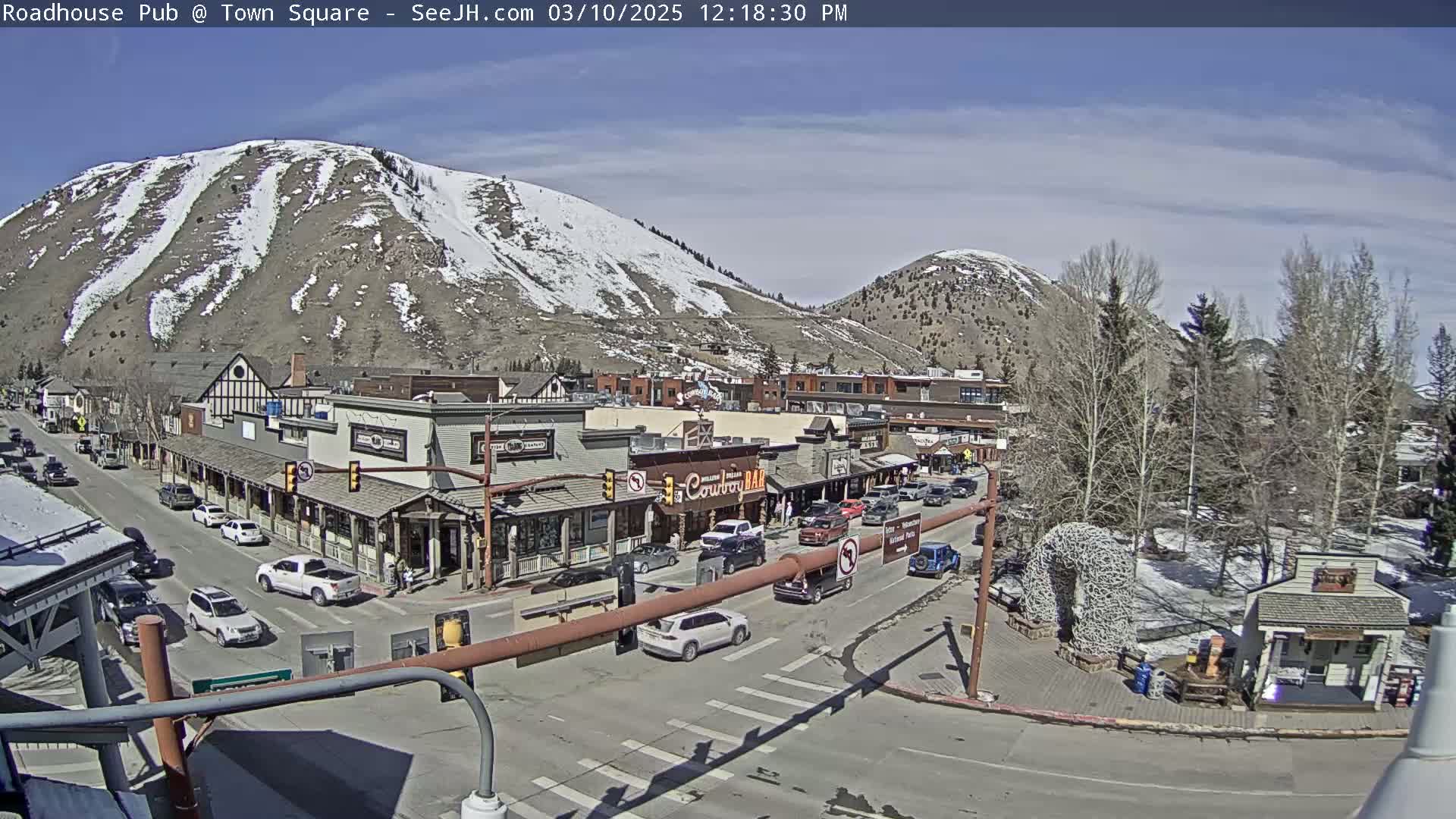A sunny day in a mountain town shows a street scene with cars and buildings, with snow visible on the surrounding hills.
