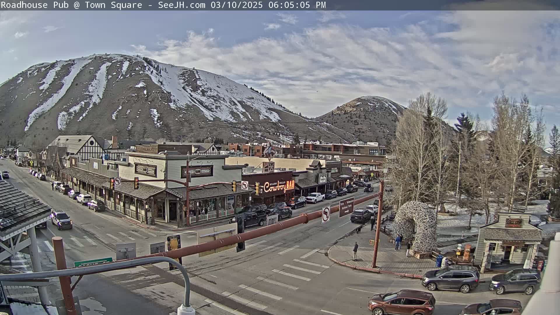 A partly cloudy afternoon scene shows a town square with cars parked and driving along streets, nestled between snow-dusted mountains.