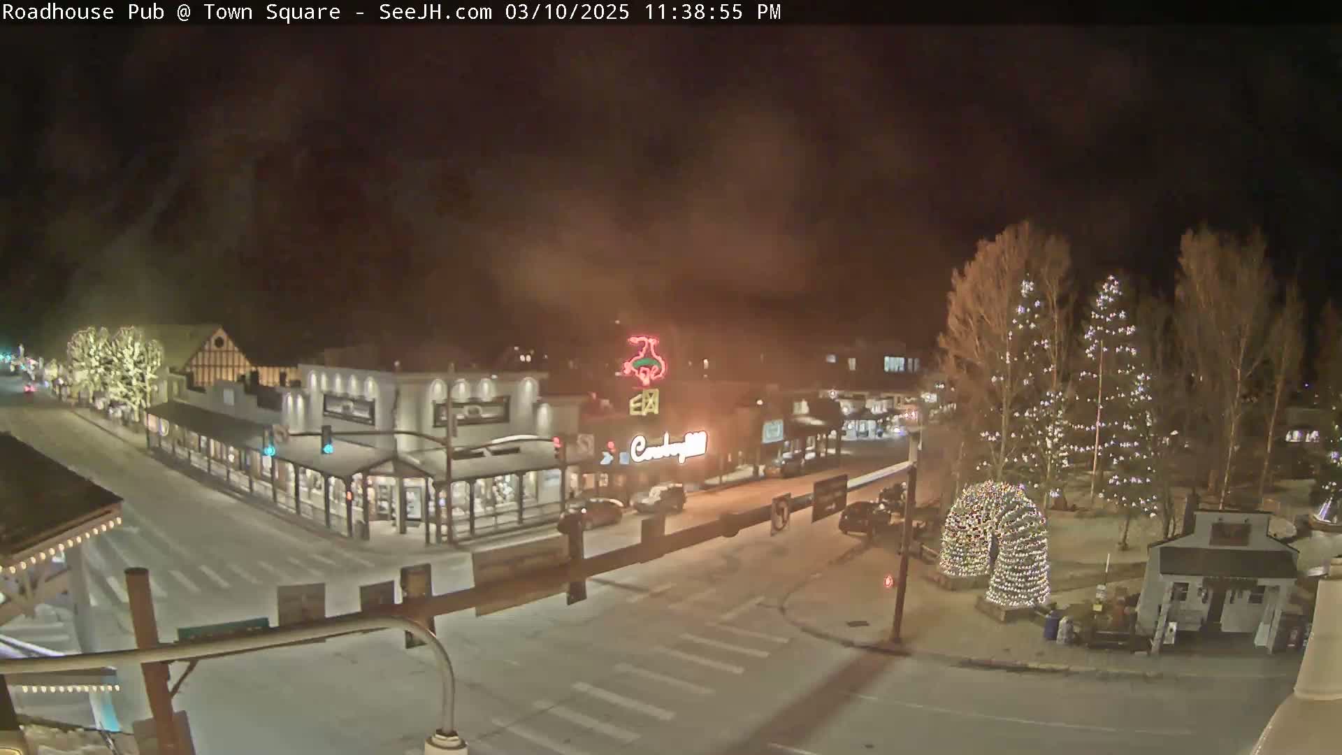 A nighttime view of a snow-covered town square with illuminated buildings and Christmas lights on trees.