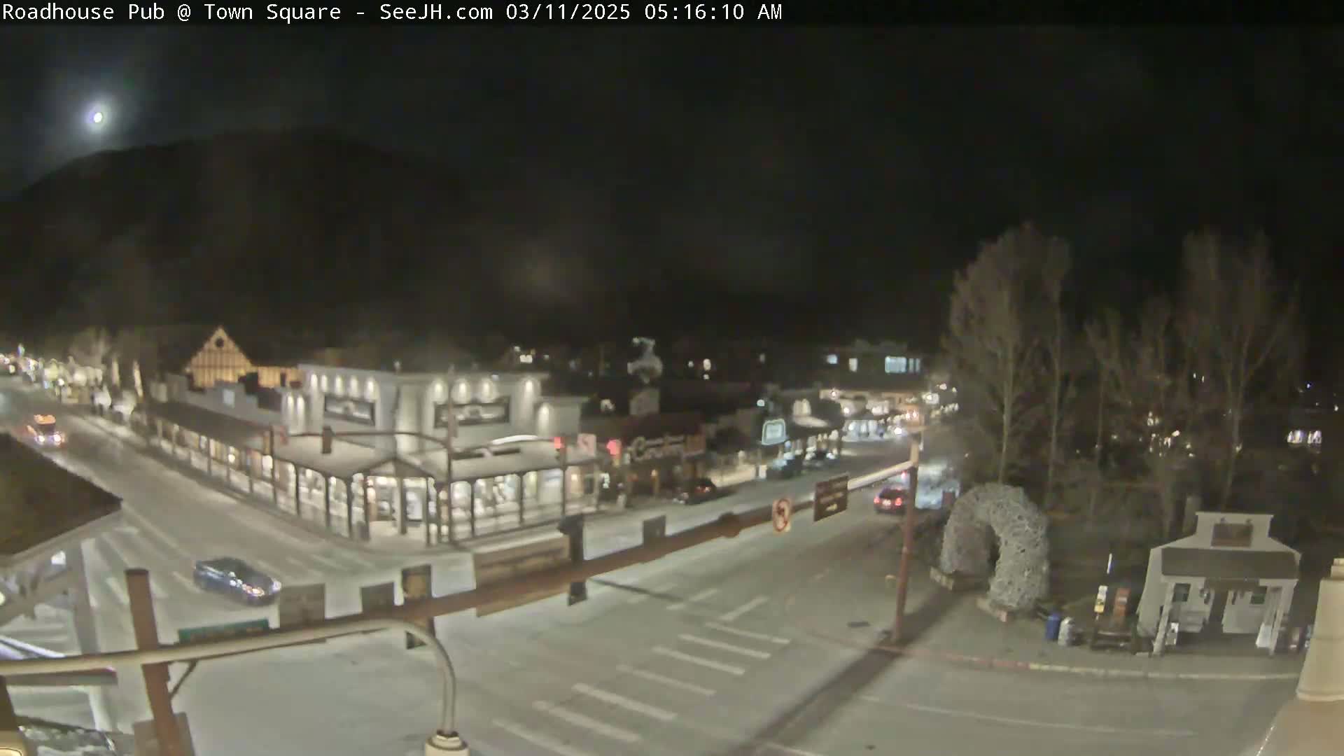 A nighttime, snow-covered town square with buildings, vehicles, and trees under a mostly clear sky with a visible moon.