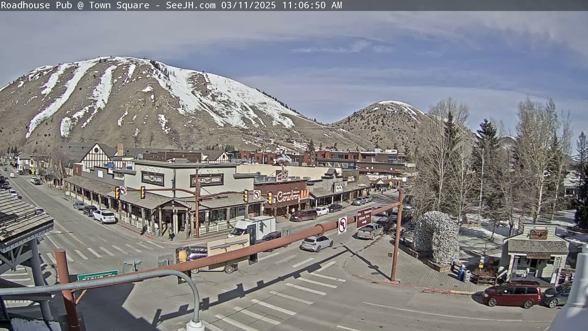 A sunny day reveals a town square with snow-dusted mountains in the background, cars parked along the streets, and buildings lining the roads.