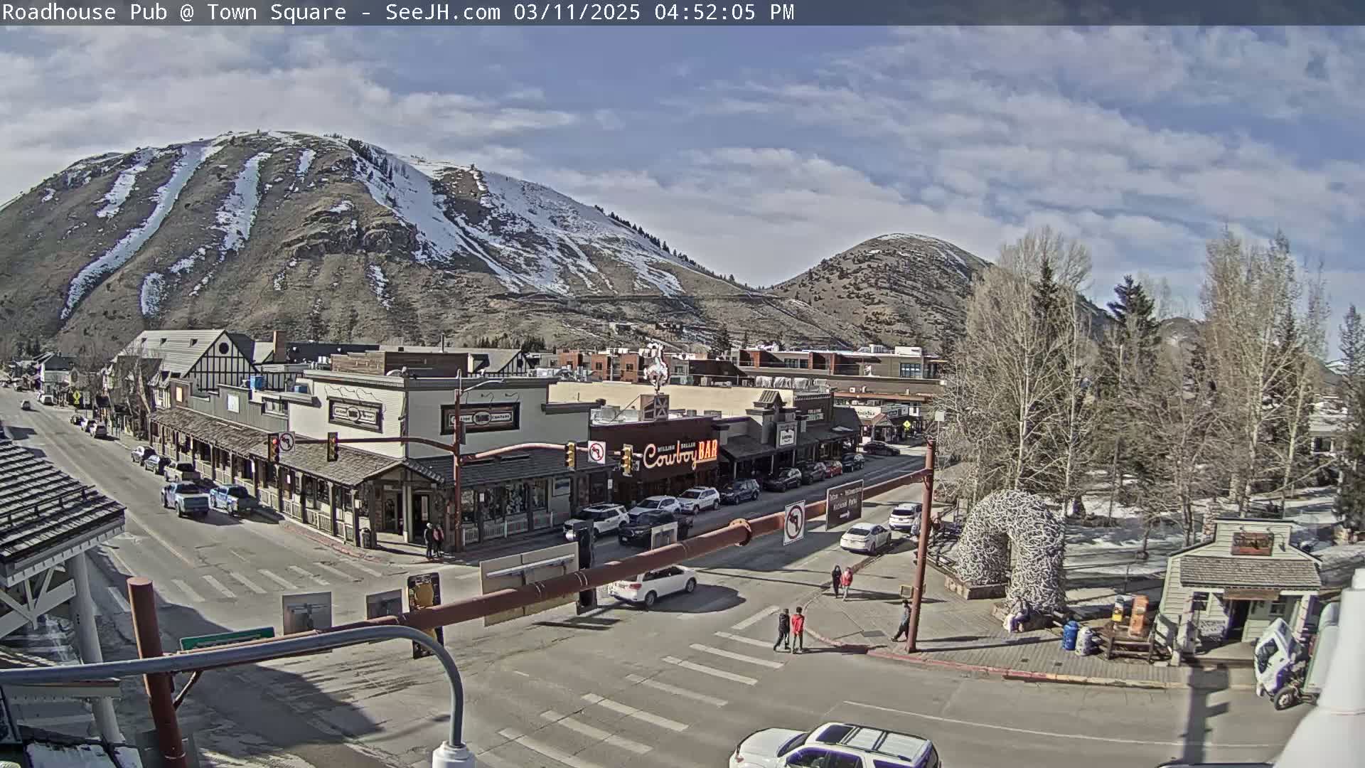 A partly cloudy day reveals a mountain town with snow-dusted mountains in the background, streets lined with buildings, and cars parked along the roadside.