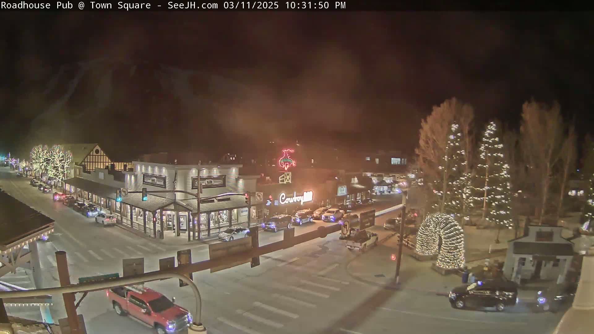A nighttime, slightly hazy view of a town square with Christmas lights illuminating buildings and trees, and several cars parked and driving on the streets.
