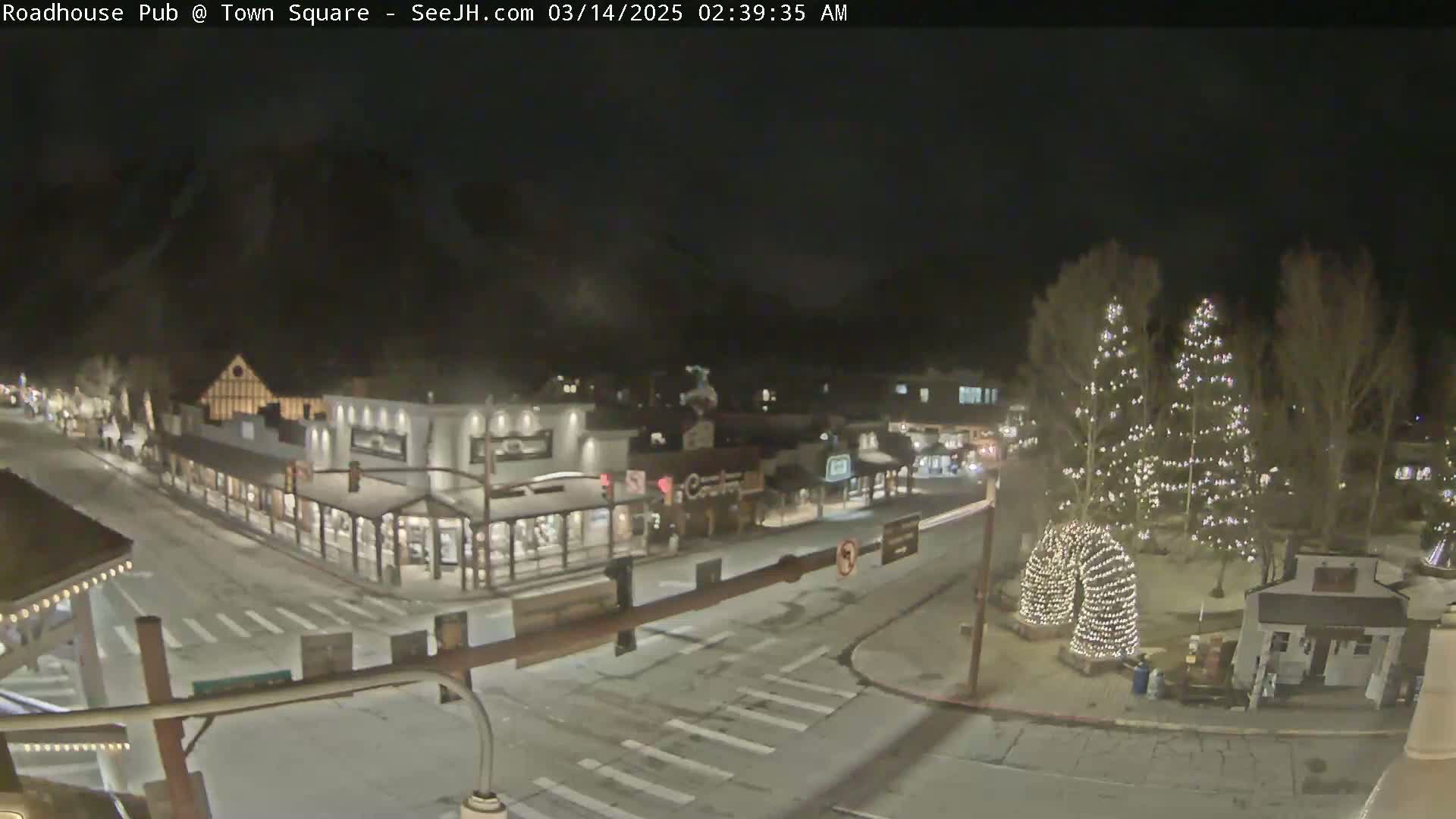 A snow-covered town square at night, illuminated by streetlights and Christmas lights on trees and structures.