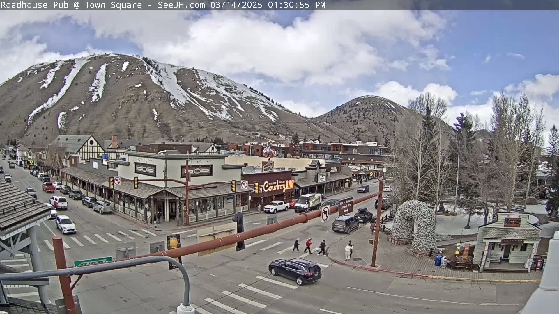 A partly cloudy day reveals a mountain town square with several cars and pedestrians, a mountain with snow patches in the background.