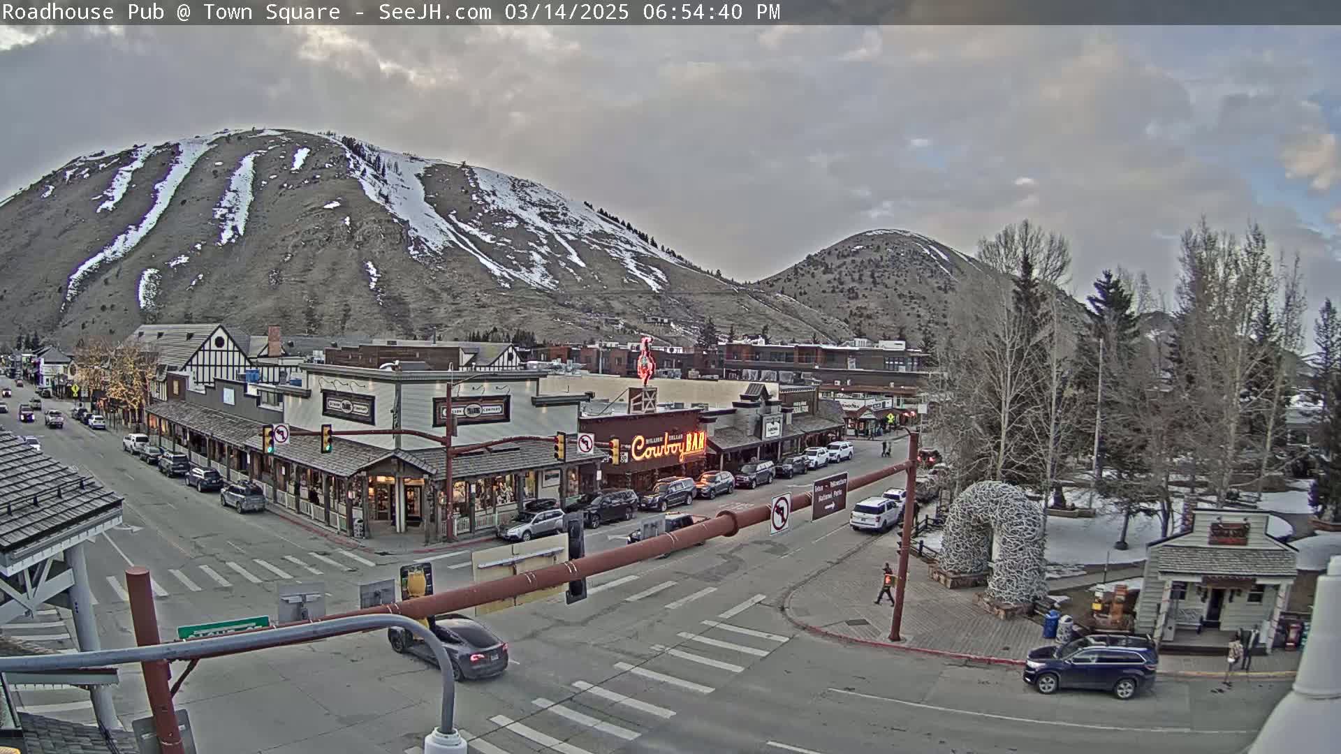 A partly cloudy afternoon scene shows a town square with shops and cars, overlooked by snow-dusted mountains.