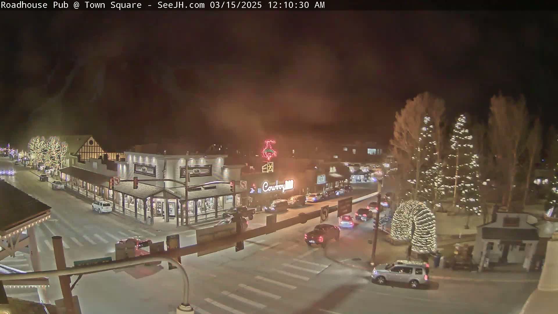 A nighttime view of a town square with several buildings, some decorated with lights, and cars driving on the roads.