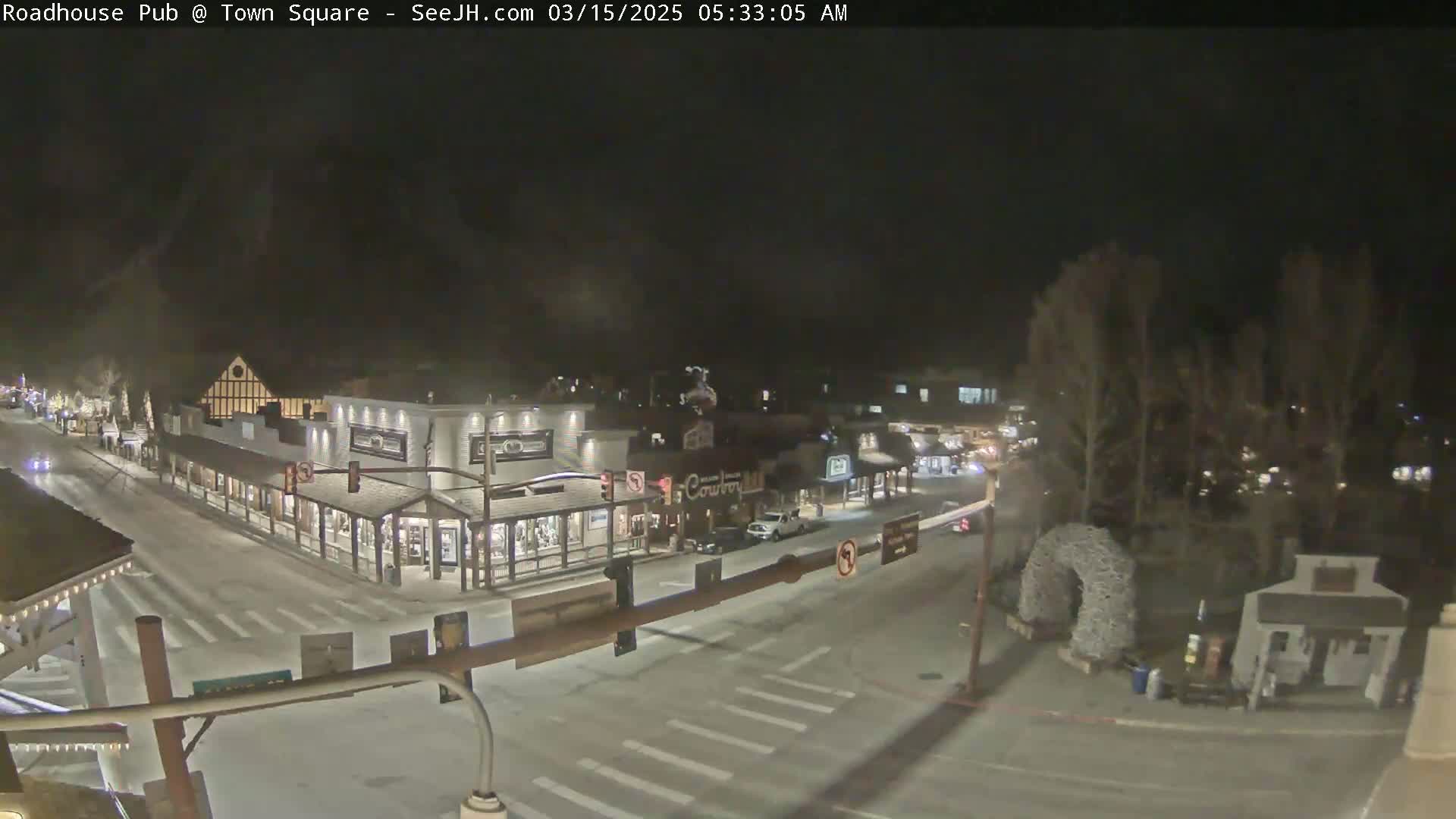 A nighttime view of a snow-covered town square with several buildings and sparsely-trafficked streets.