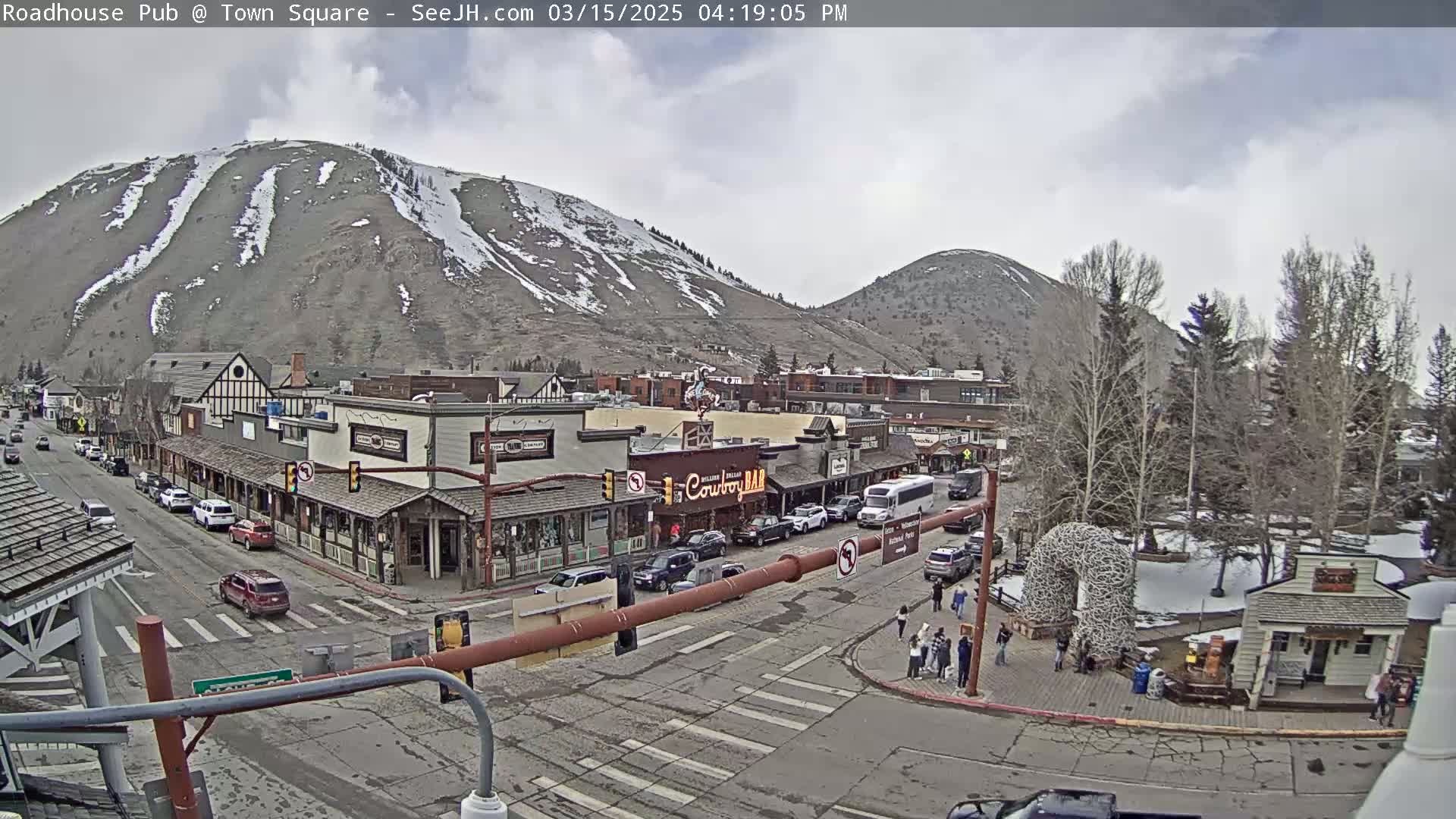 A mountain town street scene under a cloudy sky shows a main street with shops and parked cars, with snow-capped mountains in the background.