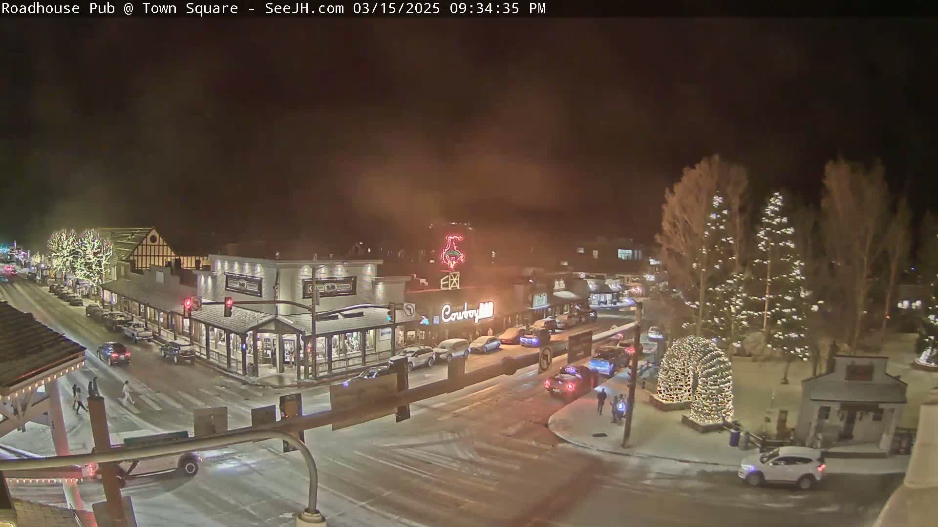 A snow-covered town square at night, illuminated by streetlights and Christmas lights on trees, with cars parked and driving along the streets.