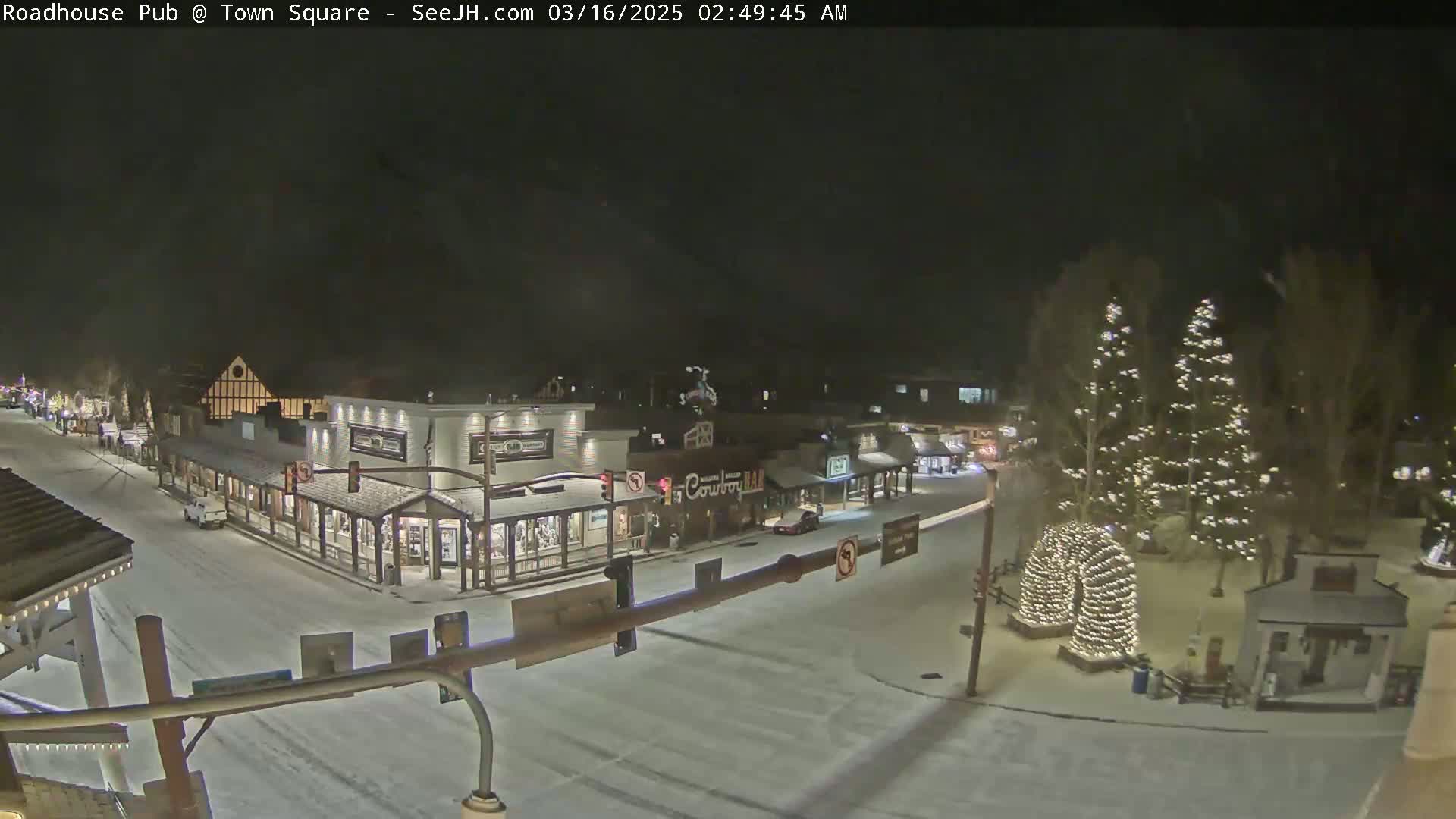 A snow-covered town square at night, illuminated by streetlights and Christmas lights on trees.