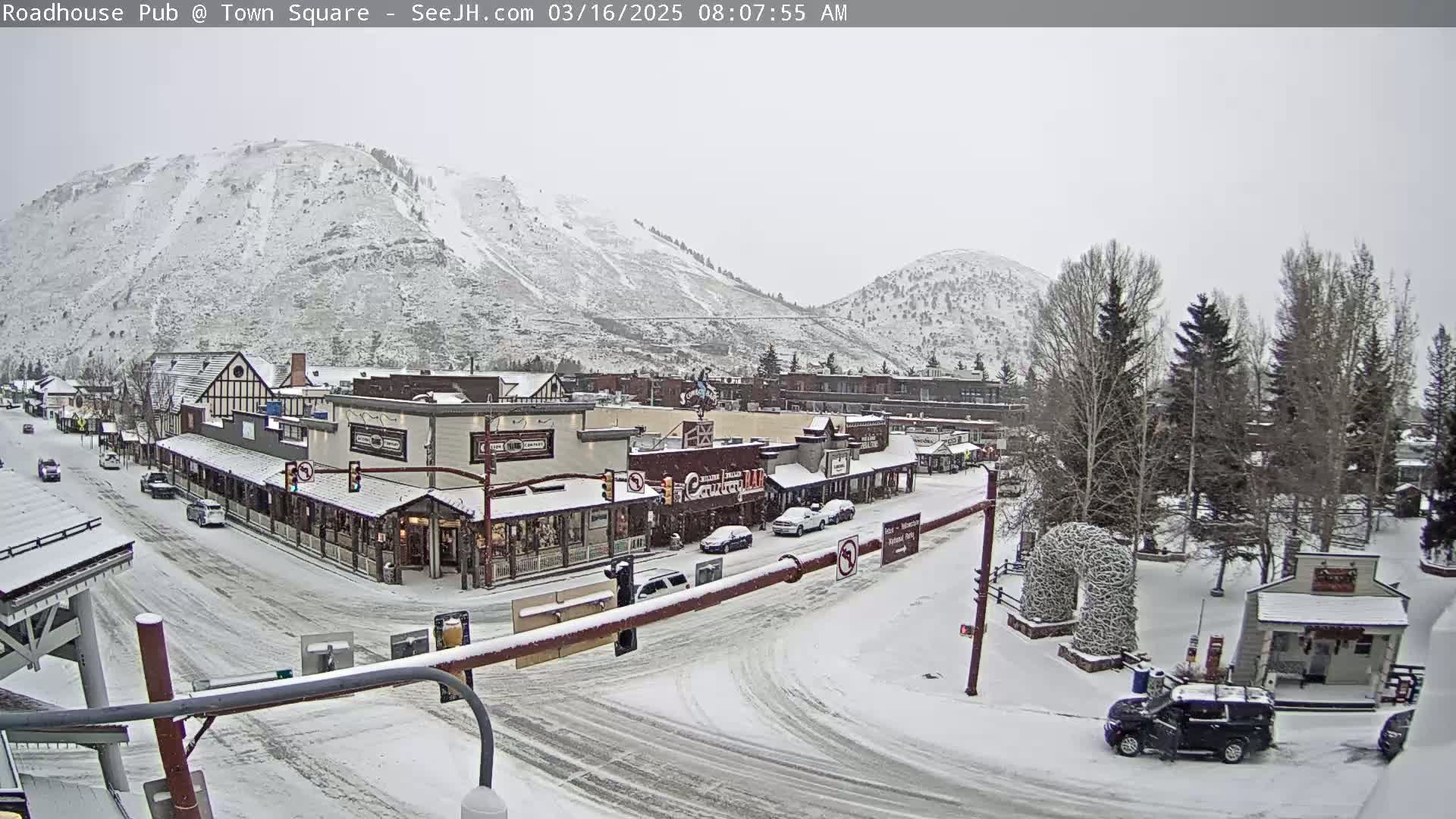 A snow-covered town square with several buildings, some cars, and trees, nestled beneath snow-dusted mountains in light snow.