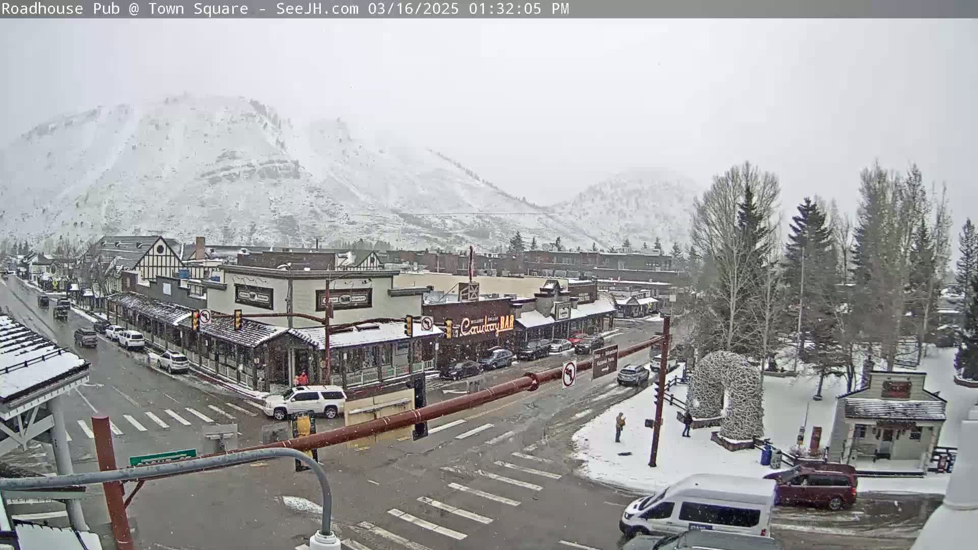 A snow-covered town square with cars and pedestrians, situated at the base of snow-dusted mountains.