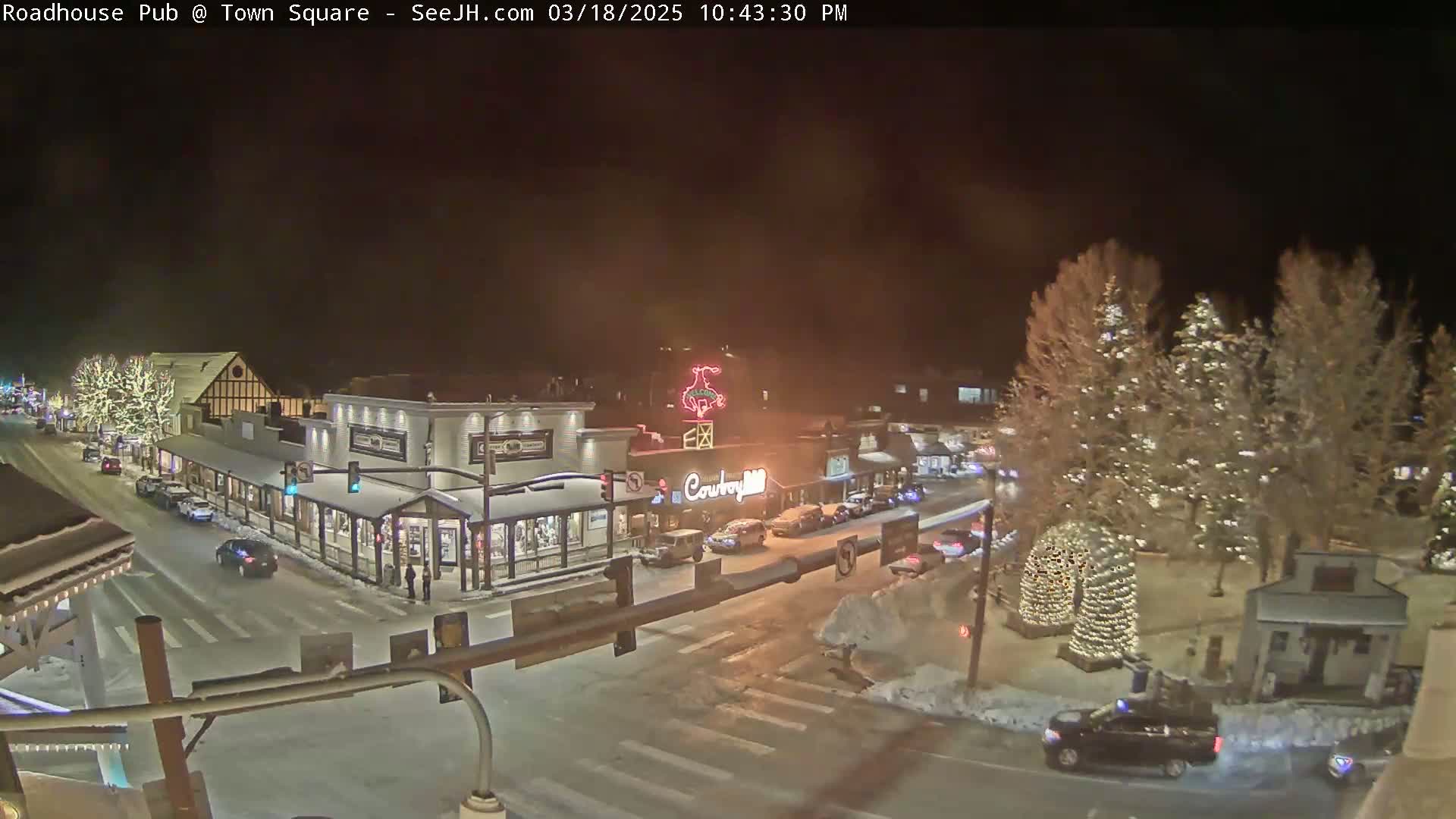 A snow-covered town square at night, illuminated by streetlights and Christmas decorations on buildings and trees, with several cars visible on the roads.