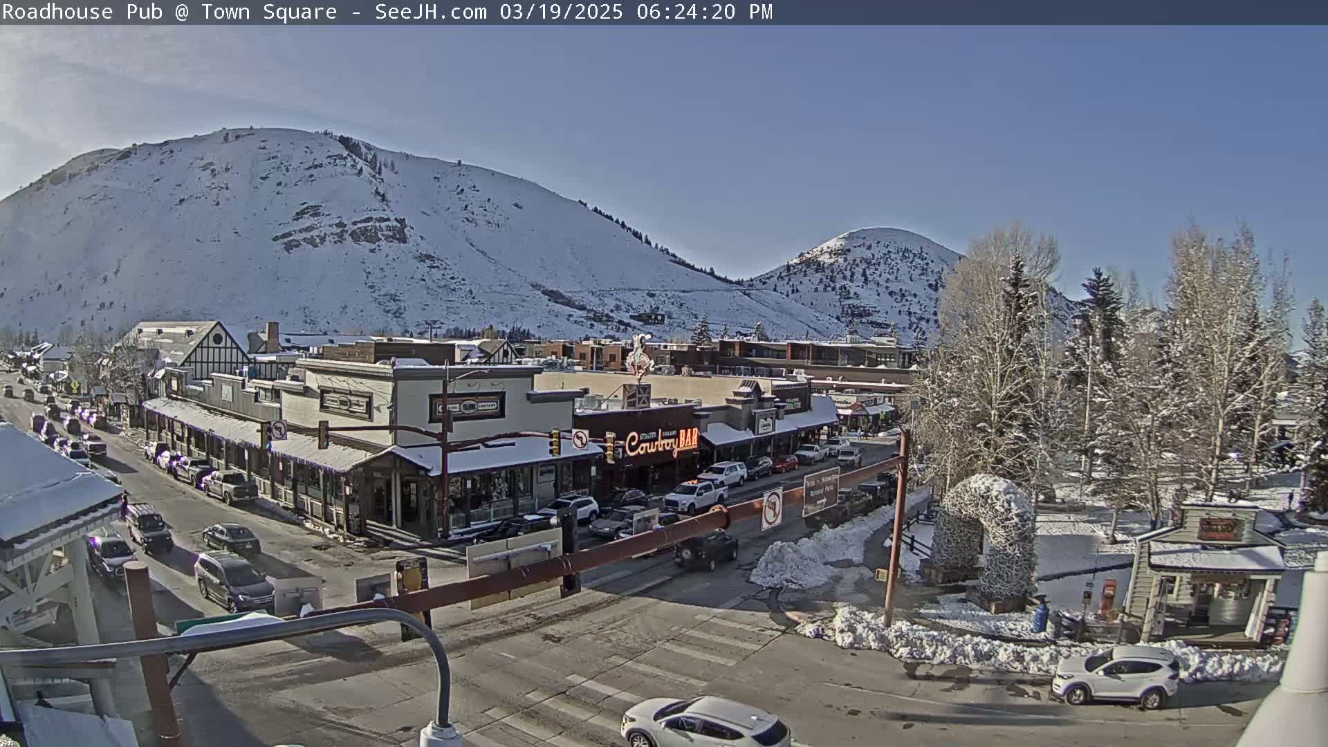 A snow-covered town nestled between snow-dusted mountains on a sunny day, with cars driving along a street lined with buildings.