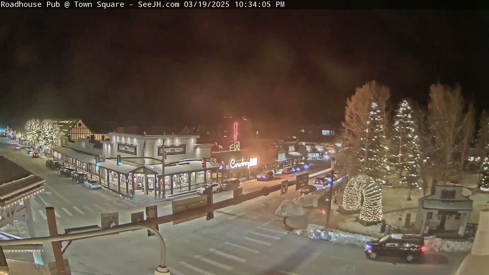 A snow-covered town square at night, illuminated by streetlights and Christmas lights on trees, shows several cars parked and driving along the streets in front of shops and restaurants.