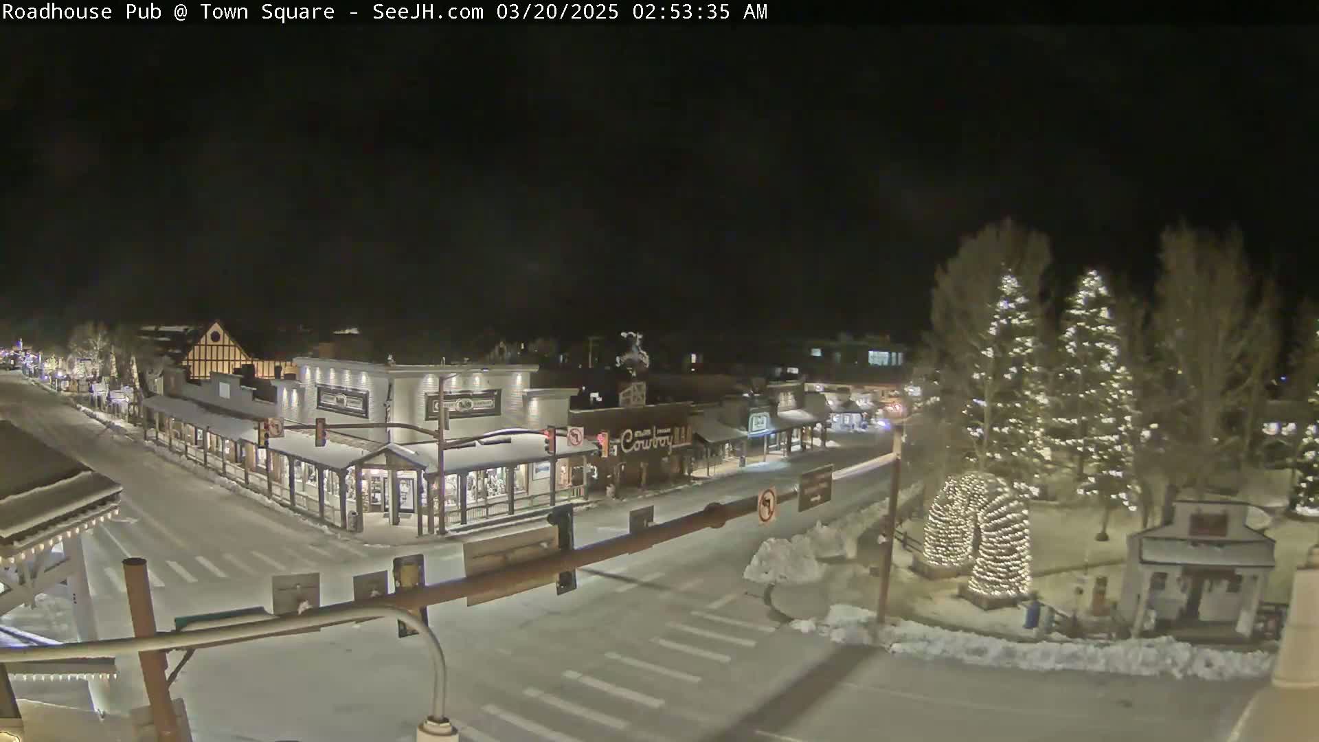 A snow-covered town square at night, illuminated by streetlights and Christmas lights on trees, shows several buildings along a street intersection.