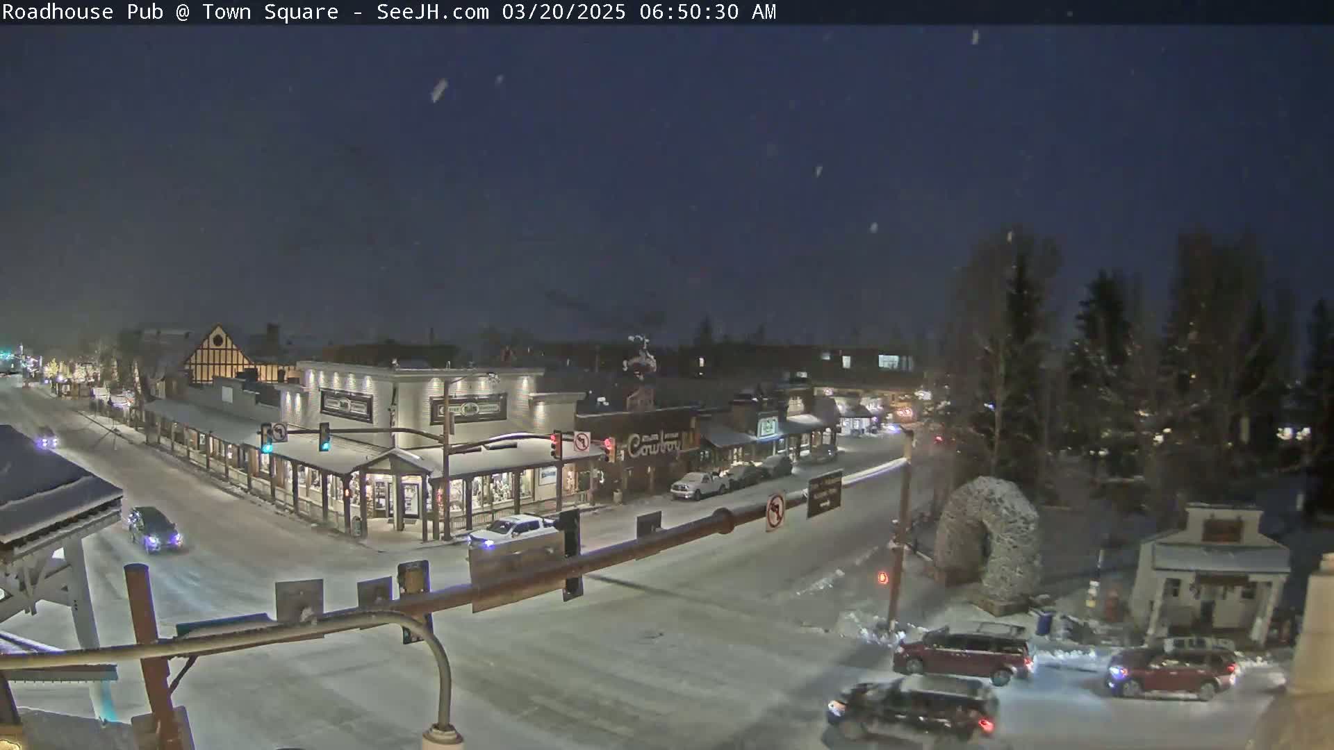 A snow-covered town square at night, with several cars on the road and buildings illuminated.