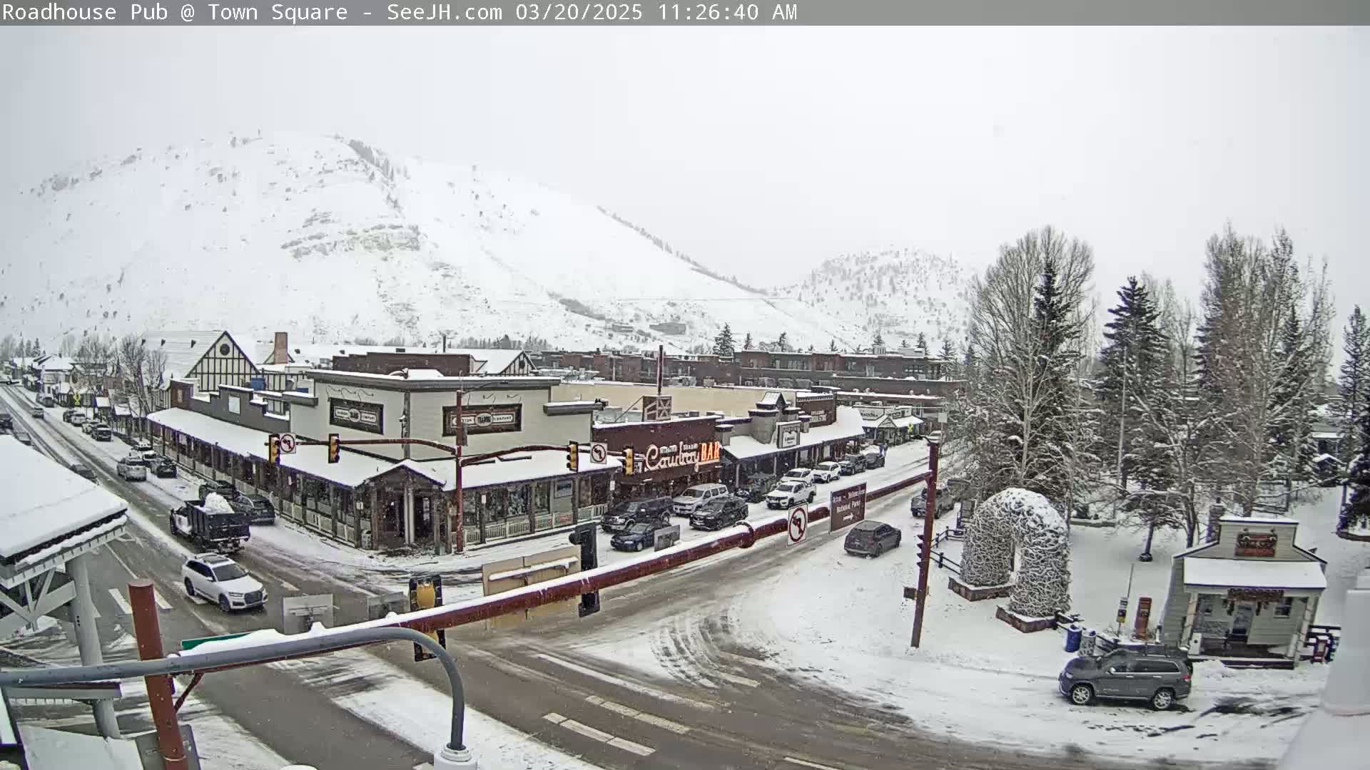 A snow-covered town square with cars parked and driving on the streets, nestled at the base of a snow-dusted mountain.