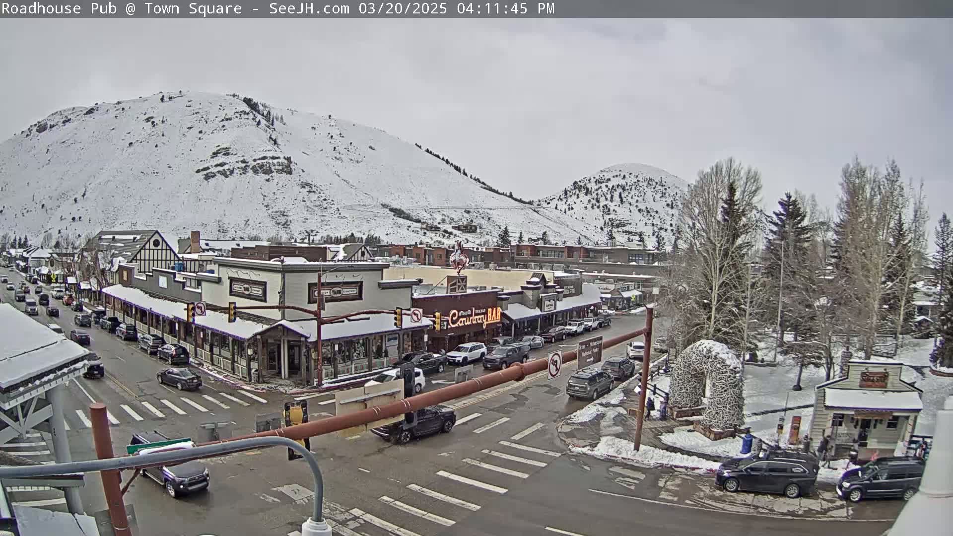 A snow-covered town square with cars parked along the street, under an overcast sky, is nestled below snow-dusted mountains.