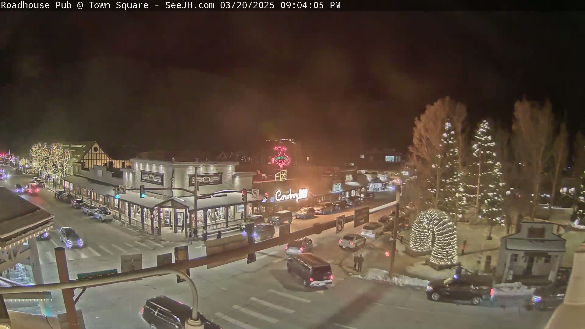 A nighttime view of a snow-covered town square with cars on the street, buildings decorated with lights, and several Christmas trees illuminated.