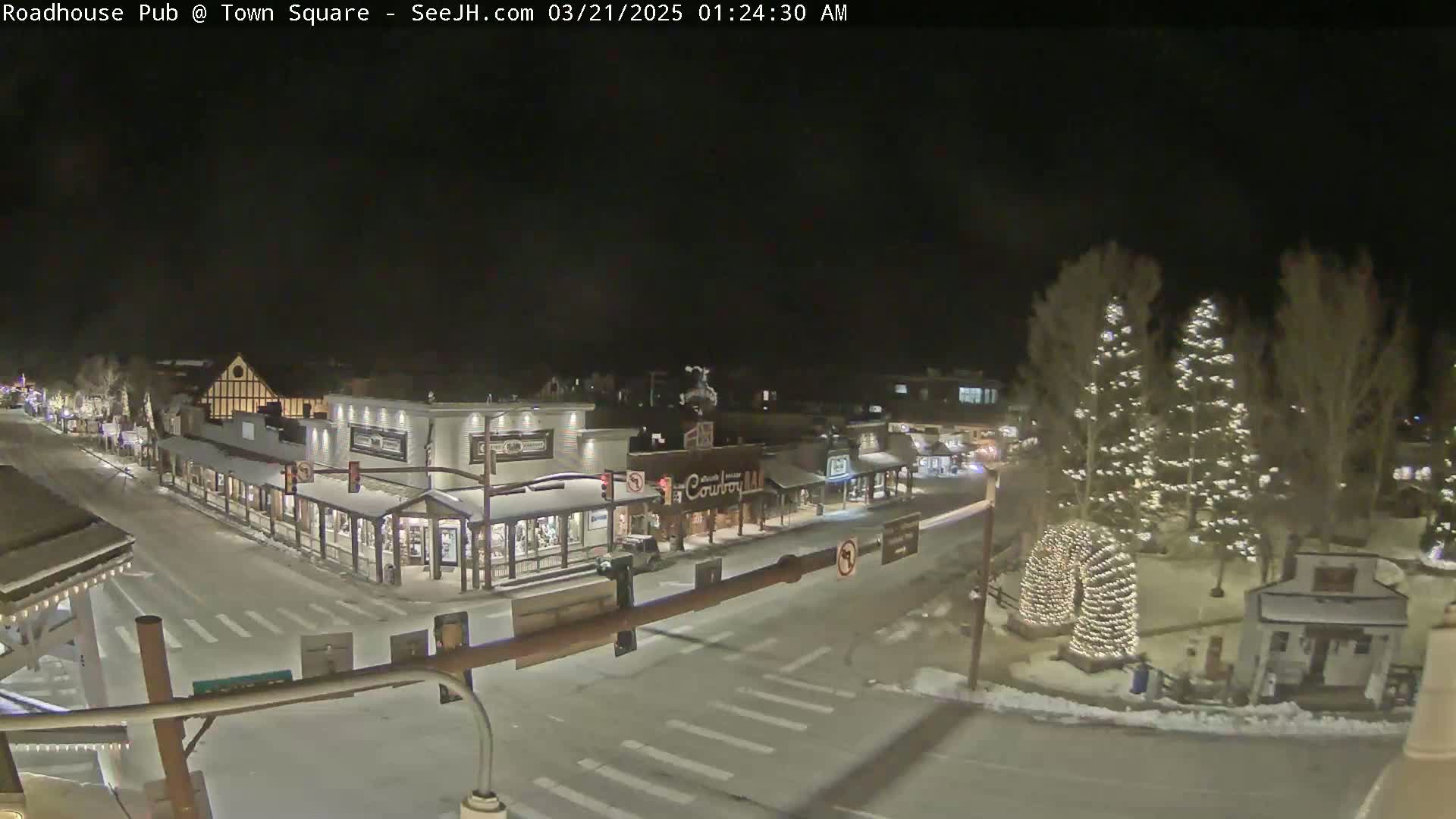 A snow-covered town square at night, illuminated by streetlights and Christmas lights on trees.