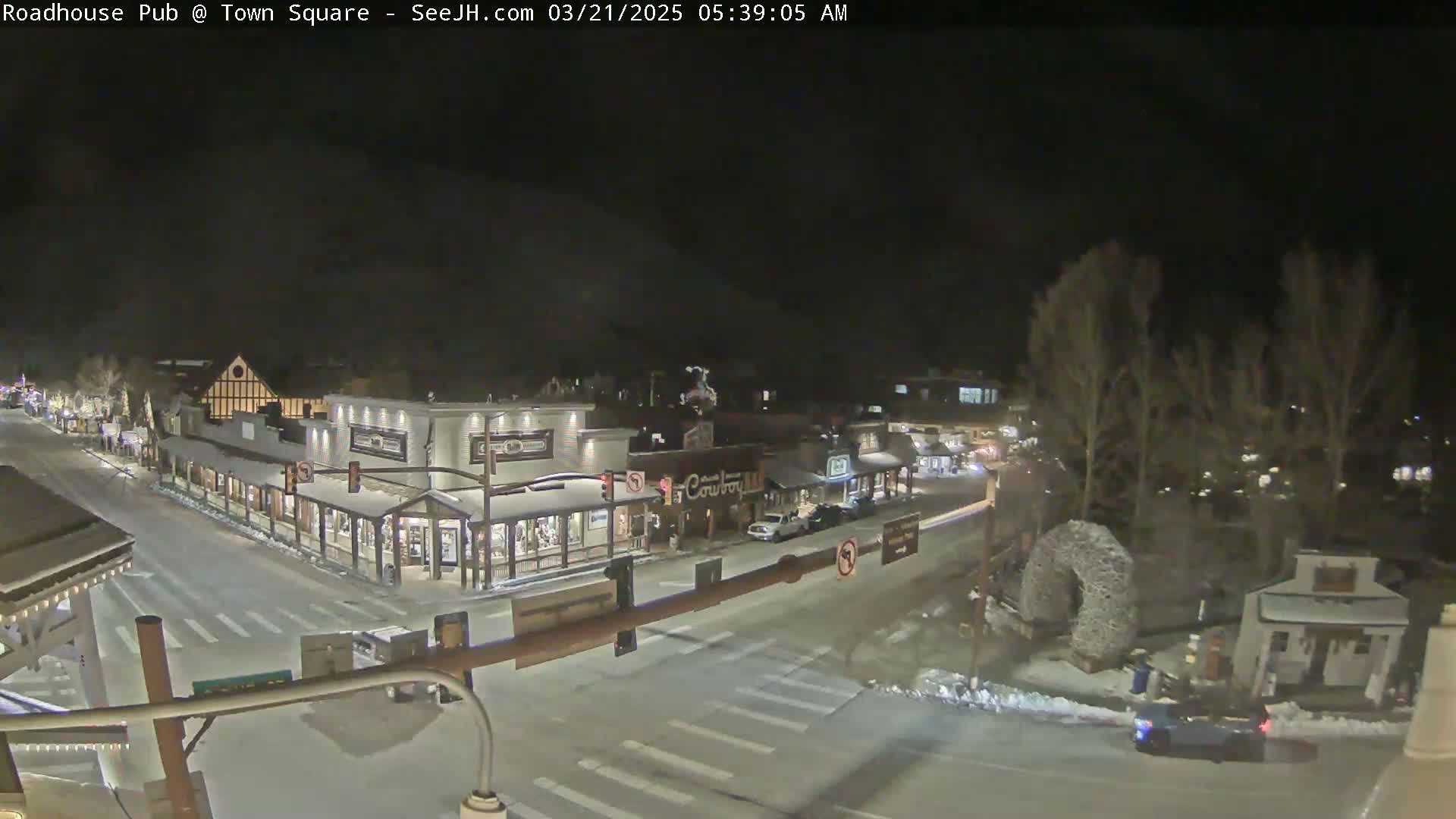 A snow-covered town square at night, with several buildings illuminated and a few vehicles visible on the roads.