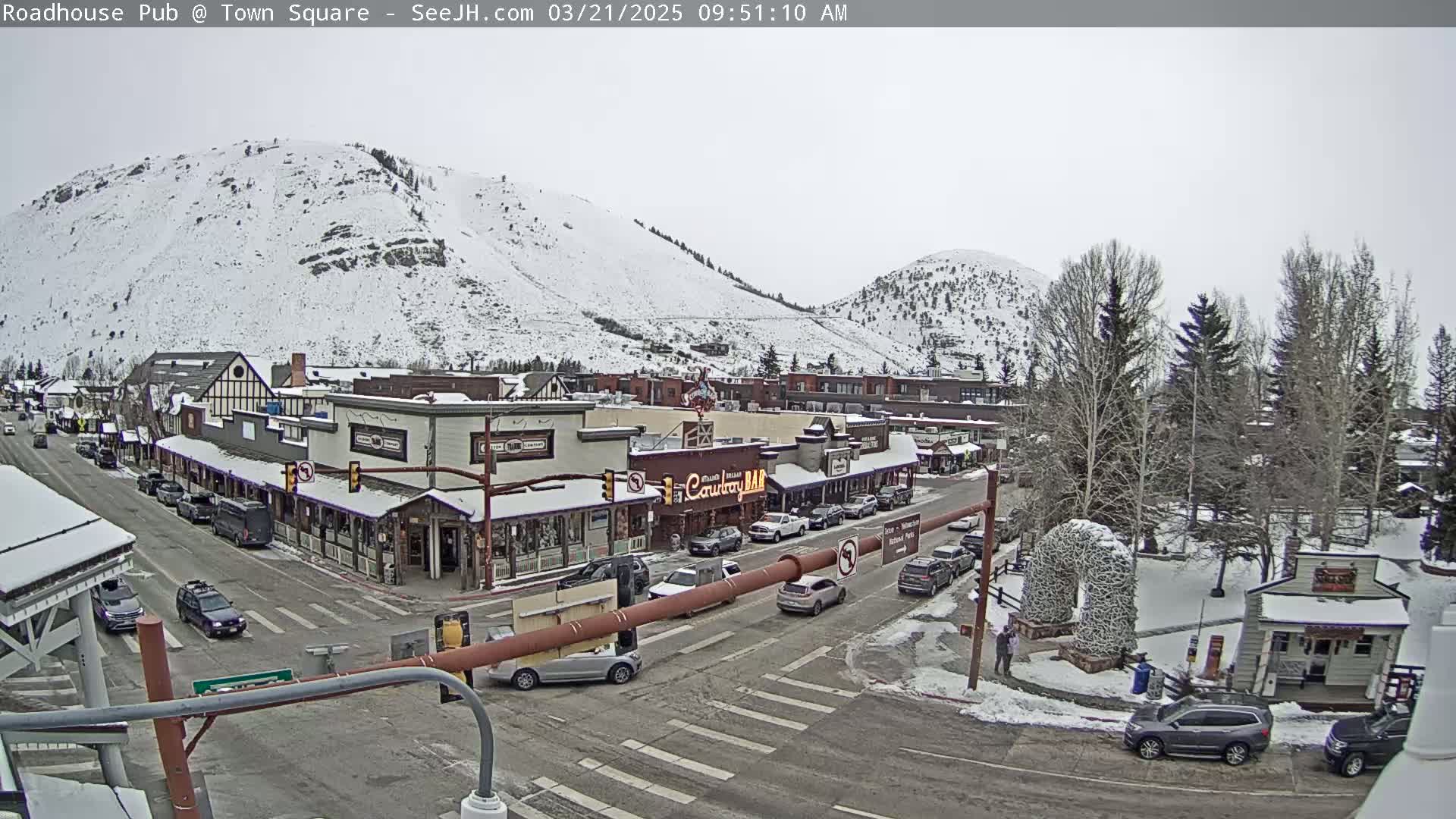 A snow-covered town square with cars parked along the street and snow-capped mountains in the background under an overcast sky.