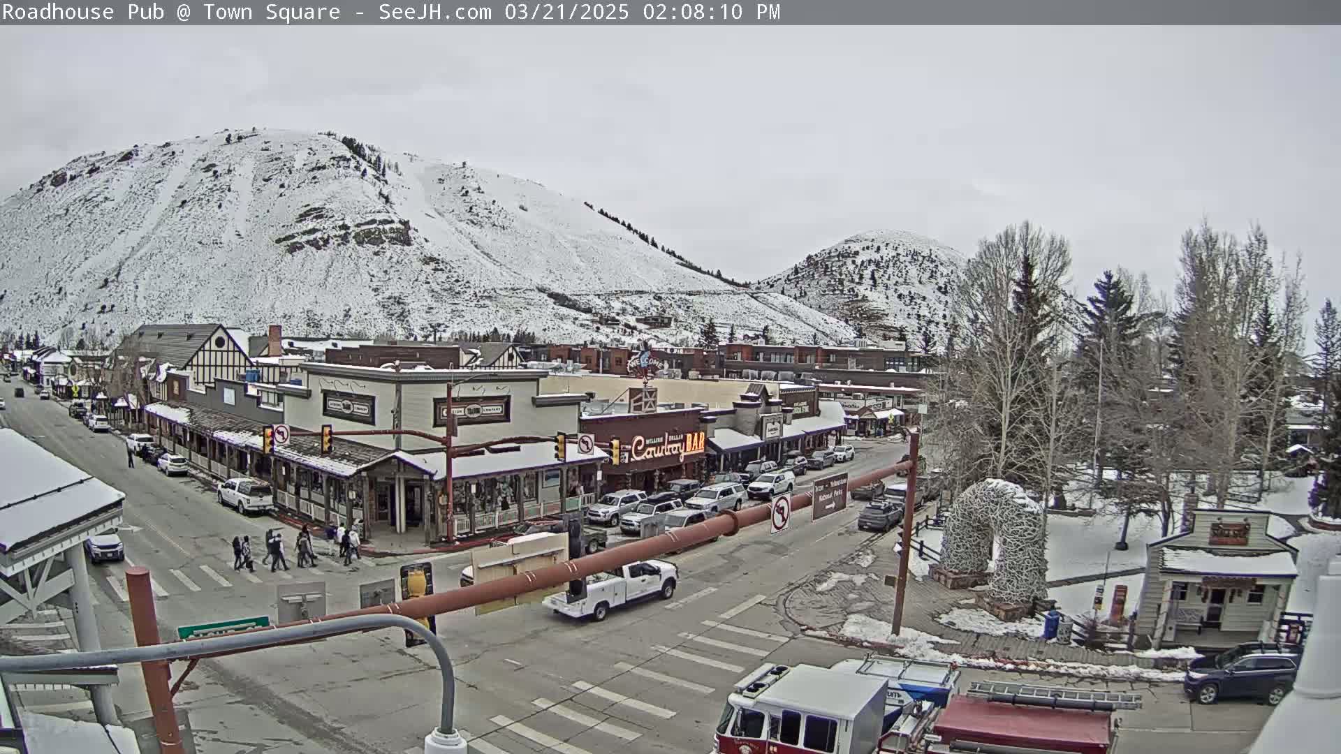 A snow-covered mountain overlooks a town square with several buildings, cars, and pedestrians on a cloudy day.