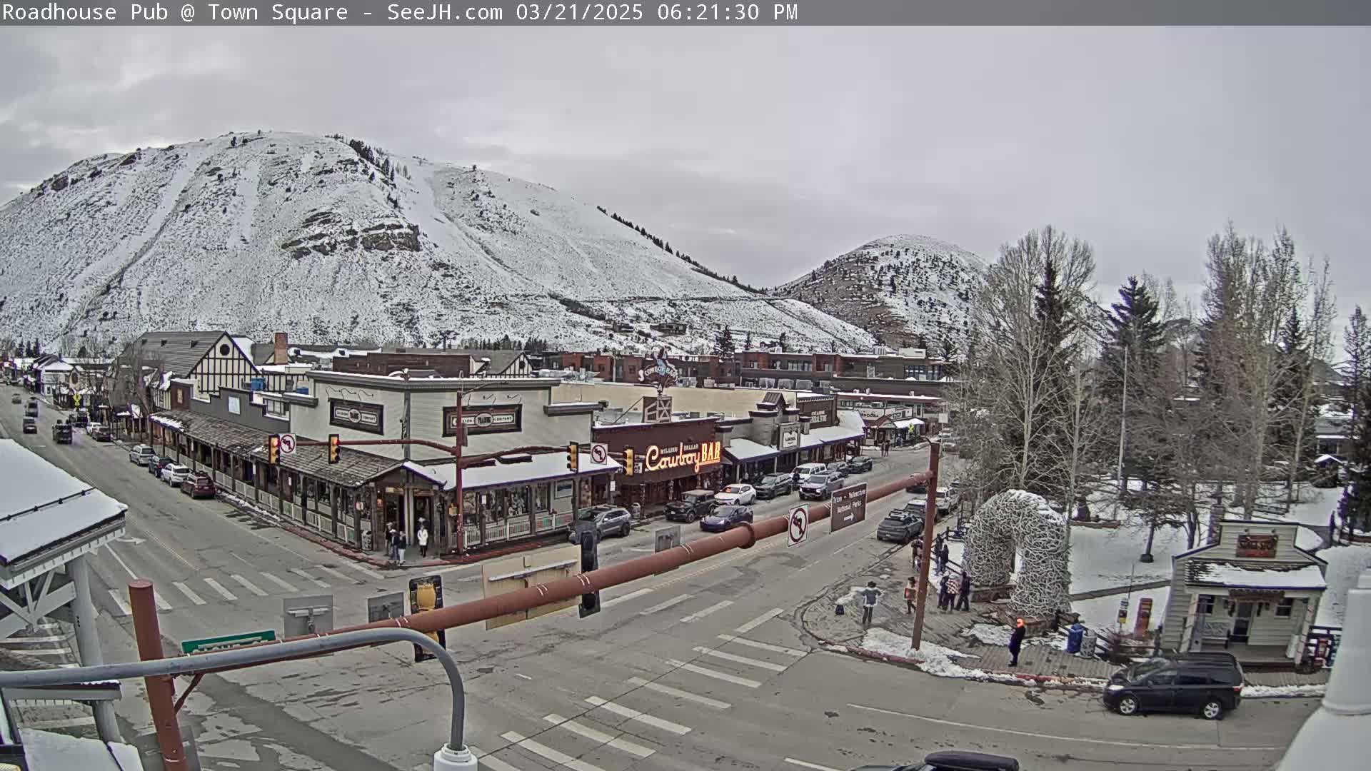 A snow-covered mountain overlooks a town square with several buildings and cars, under an overcast sky.