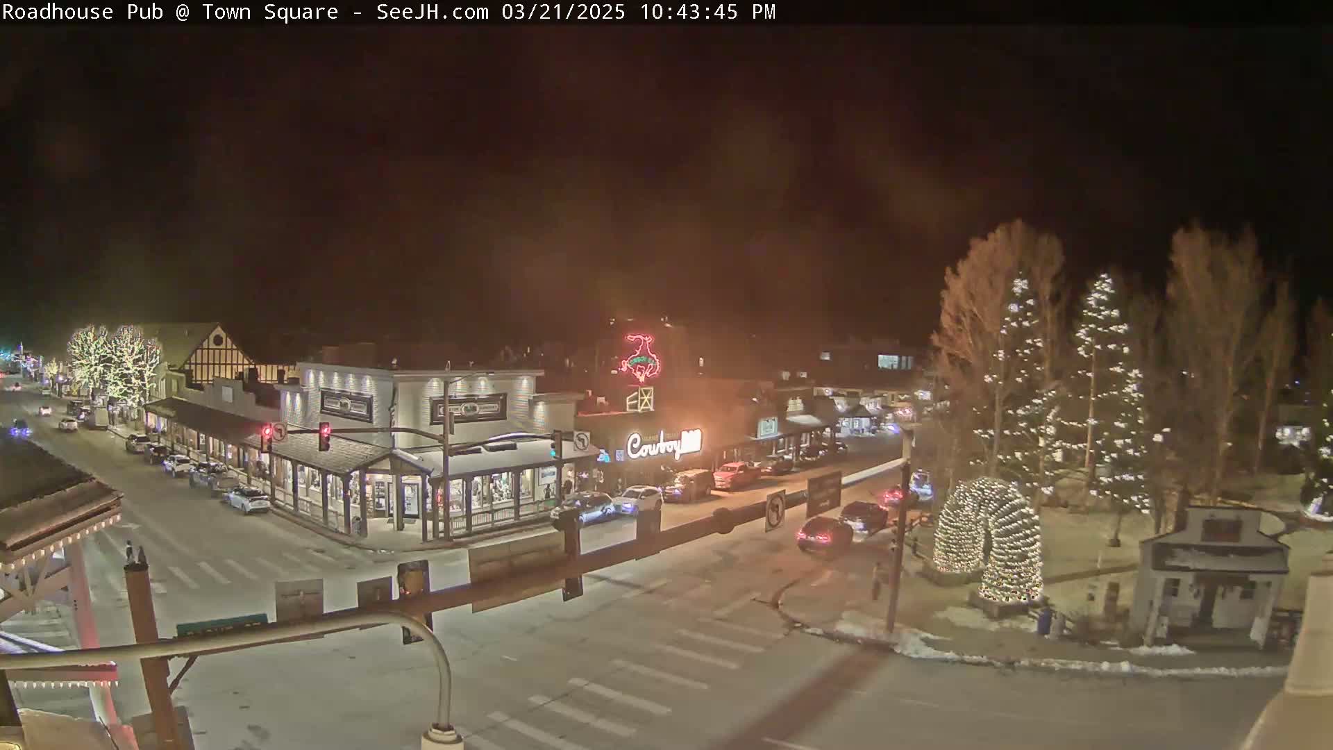 A nighttime, overhead view of a town square with several buildings and decorated trees, some cars on the road, and light snow on the ground.