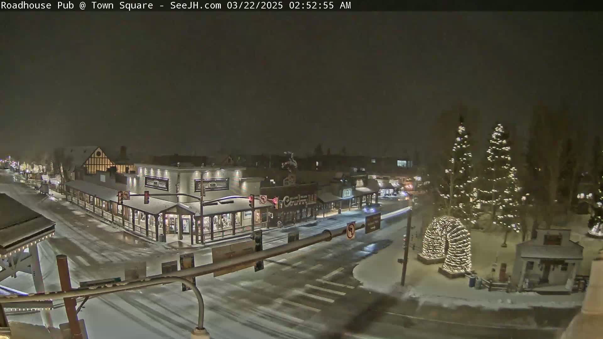 A snow-covered town square at night, illuminated by streetlights and Christmas lights on trees, with cars driving on snow-covered roads.