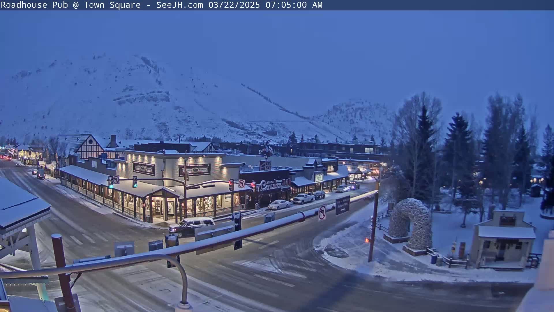 A snow-covered town square at twilight, with cars parked along the street and a snow-covered mountain in the background.