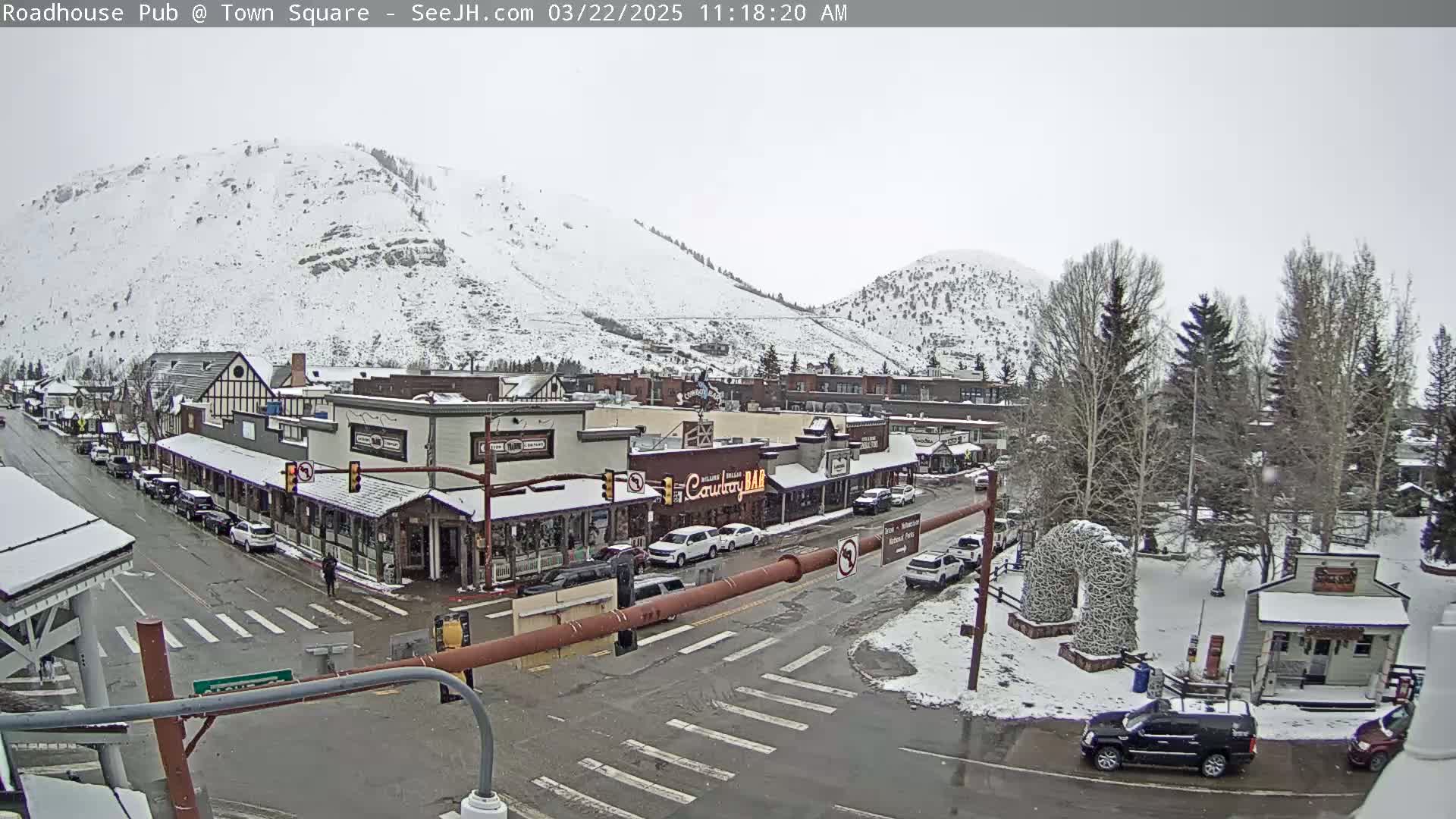 A snow-covered town square with buildings, cars, and trees, situated below snow-dusted mountains under an overcast sky.